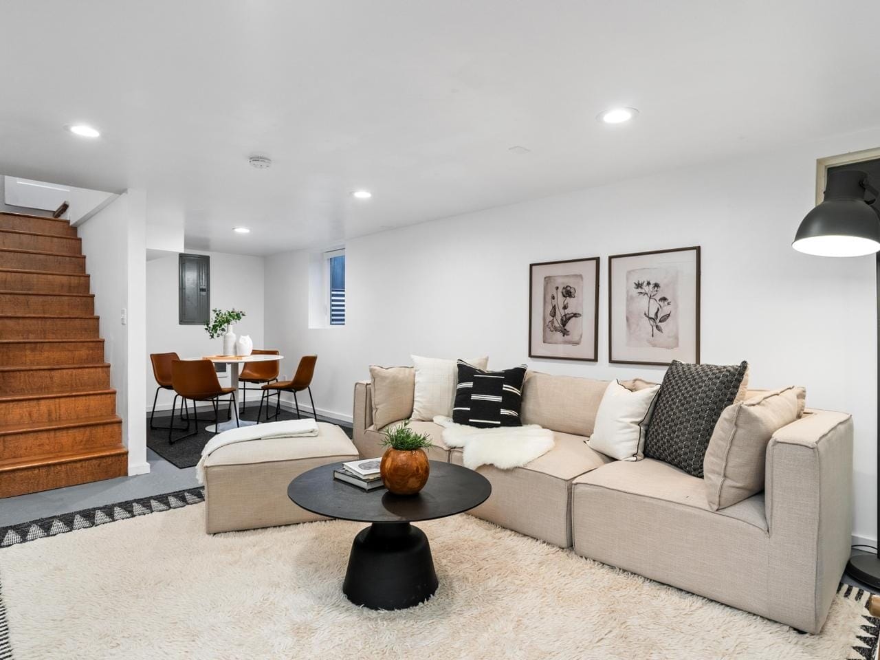 Modern living room with a beige sectional sofa, black coffee table, and decorative cushions. Framed art hangs above the couch. A dining area with a round table and chairs is visible. Wooden stairs lead up, and the room is well-lit with recessed lights.