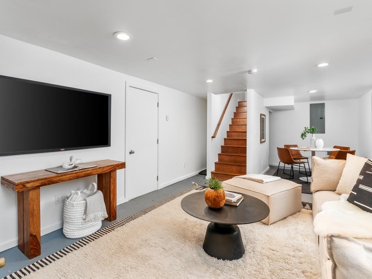 A modern living room with a wall-mounted TV, a beige sofa, and a black coffee table. Wooden stairs lead upwards. A dining area with a round table and chairs is visible in the background. The decor is minimal with neutral tones and a small plant on the table.