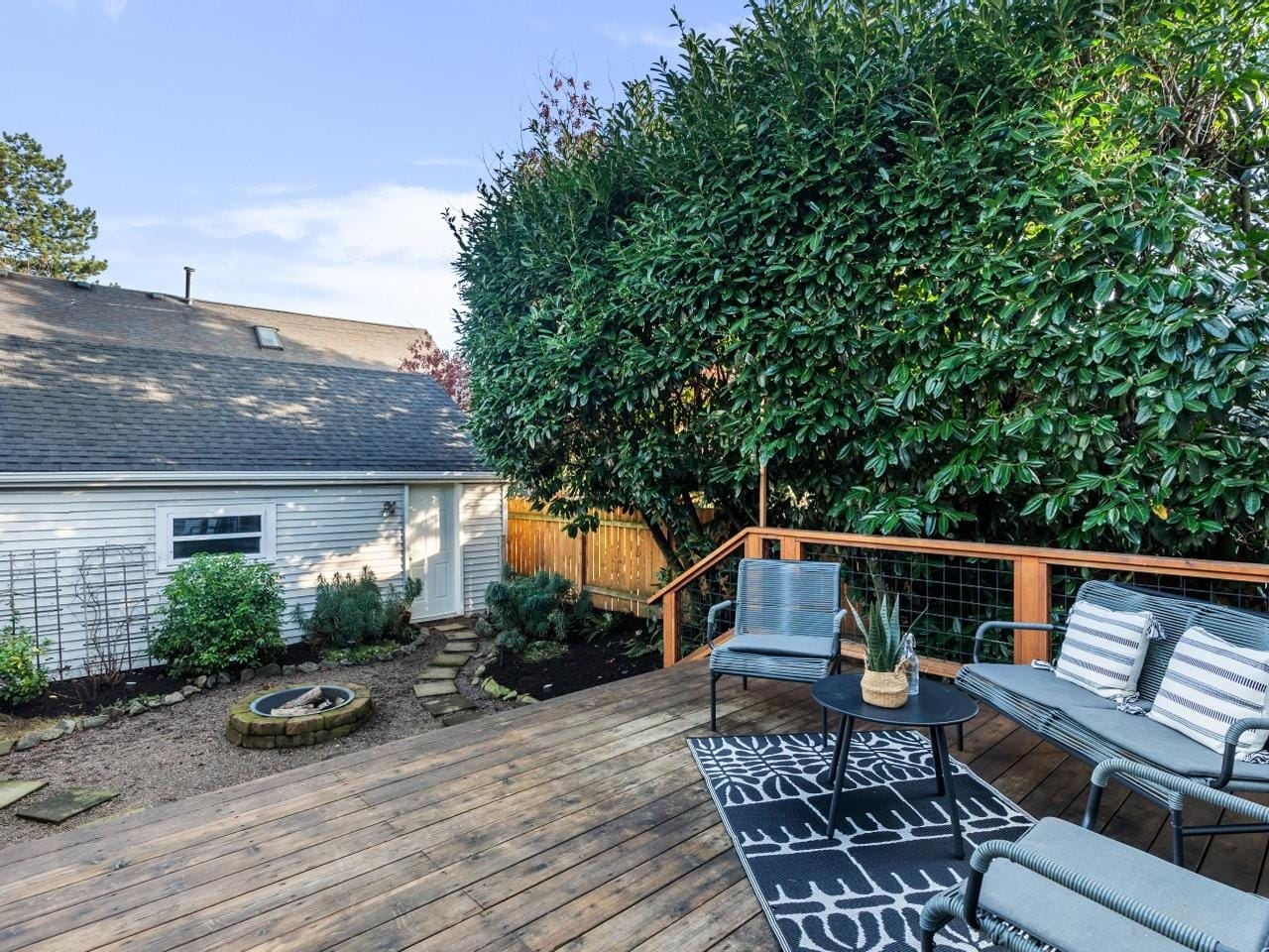 A wooden deck with striped patio furniture and a small table. A rug is placed under the table, and theres a large leafy bush nearby. In the background is a small house, a stone fire pit, and a dirt path. The sky is clear and blue.