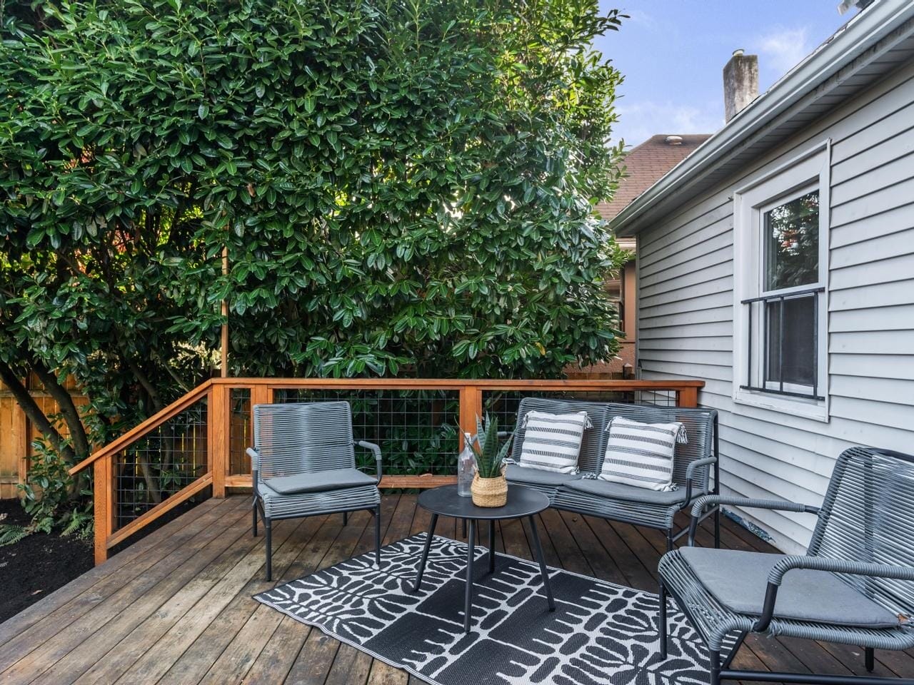 A cozy outdoor deck with two gray chairs and a matching loveseat around a small round table. A patterned black and white rug covers part of the wooden floor. Lush green bushes border the deck, and the side of a house is visible.