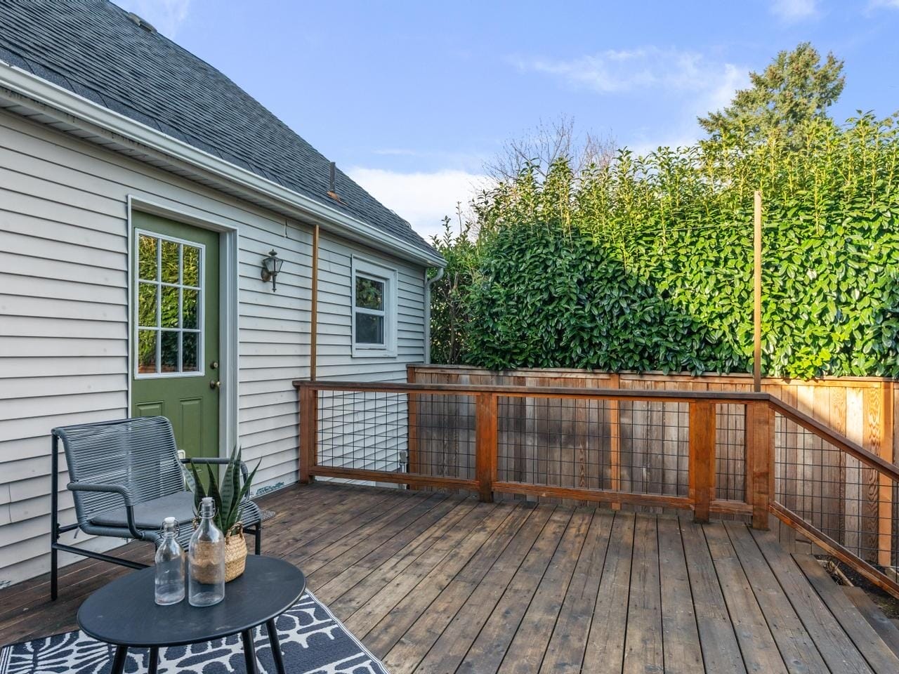 A cozy wooden deck with a small table and two chairs. The table has two clear bottles and a small plant. Greenery and a tall hedge surround the area, and theres a house with a green door and gray siding in the background.