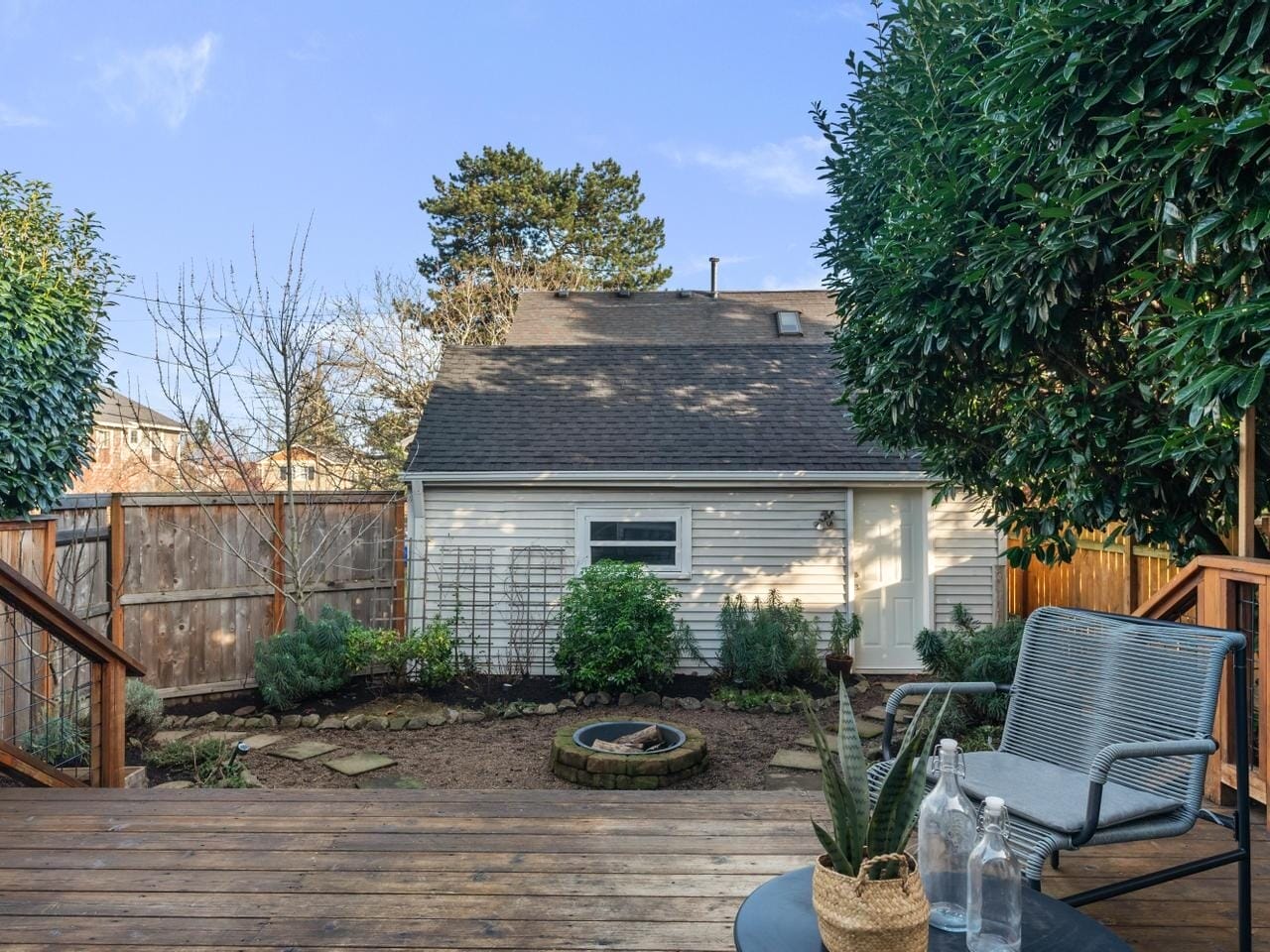 A small backyard with a wooden deck features a gray chair and a round table with plants. A garden with shrubs and a circular stone fire pit is in front, with a white shed bordered by wooden fences and lush greenery in the background.