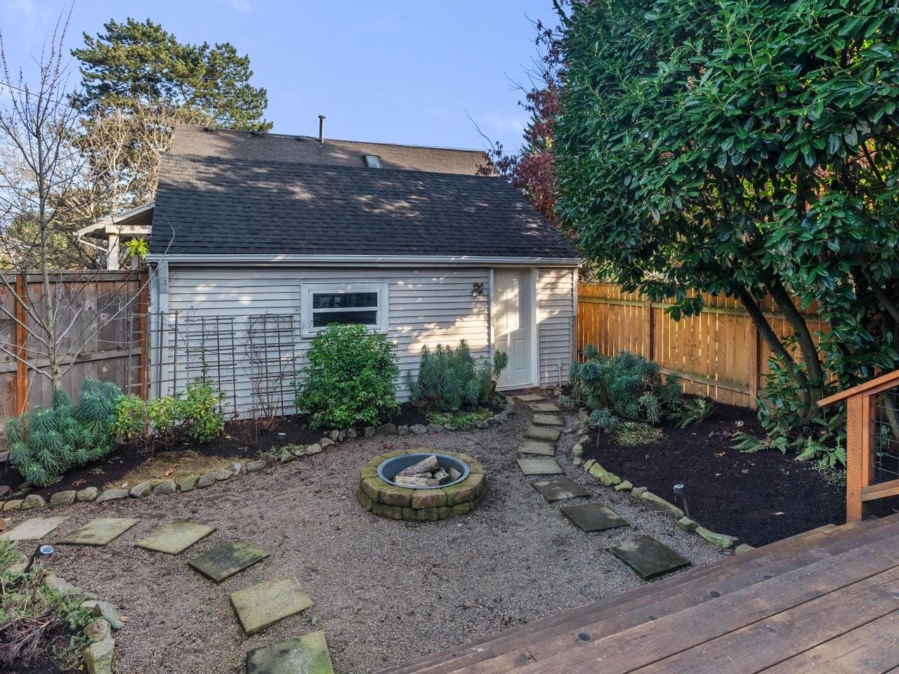 A backyard garden featuring a gravel pathway with stone steps leading to a circular fire pit. The space is bordered by a wooden fence and various shrubs, with a small white shed in the background under a clear blue sky.