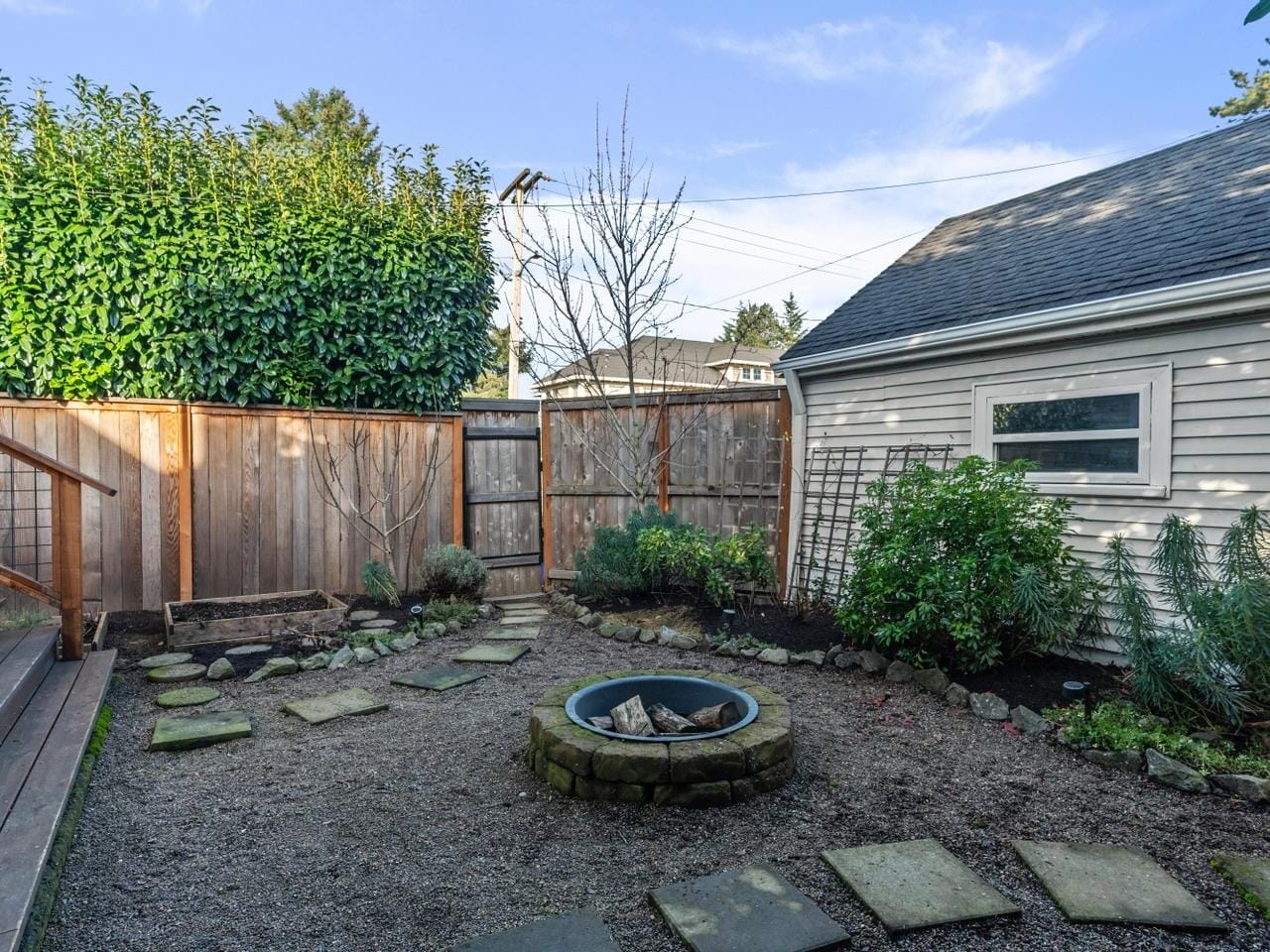 A backyard with a gravel-covered ground, stone pathway, and a circular stone fire pit. It is enclosed by a wooden fence and features shrubs and a tree. A small building with a window is on the right, and stairs lead up to a wooden deck on the left.