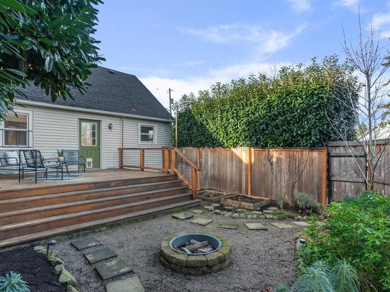 A backyard with a wooden deck featuring patio furniture. Theres a fire pit on a gravel area surrounded by stepping stones. Wooden fencing encloses the yard, with tall hedges providing greenery and privacy. A small tree is planted near the fence.