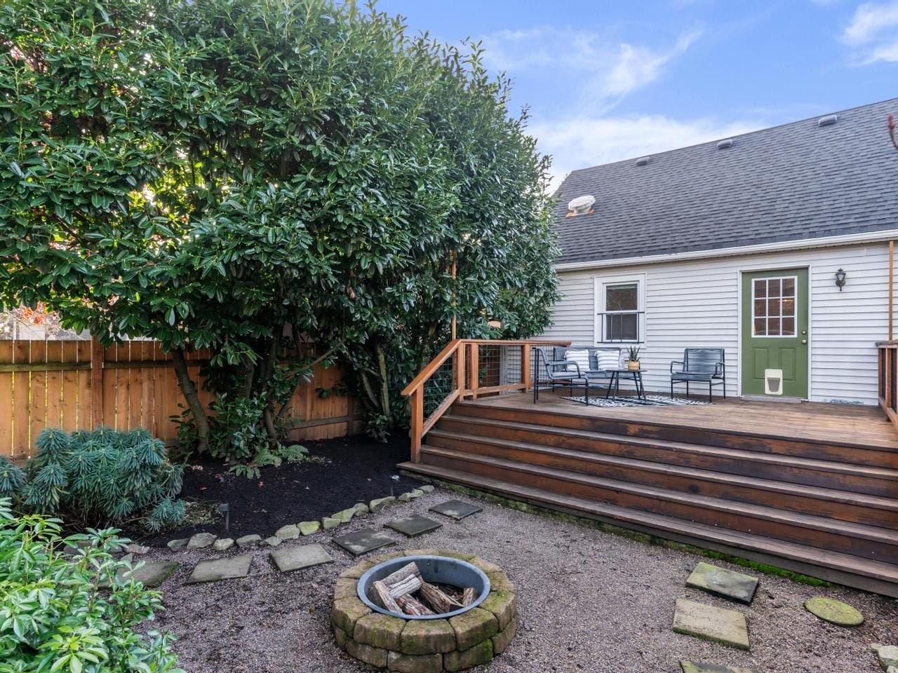 A backyard features a wooden deck with patio furniture, a small fire pit with stacked wood, and a gravel area. A large tree and a wooden fence frame the space, and the side of a white house with a green door is visible.