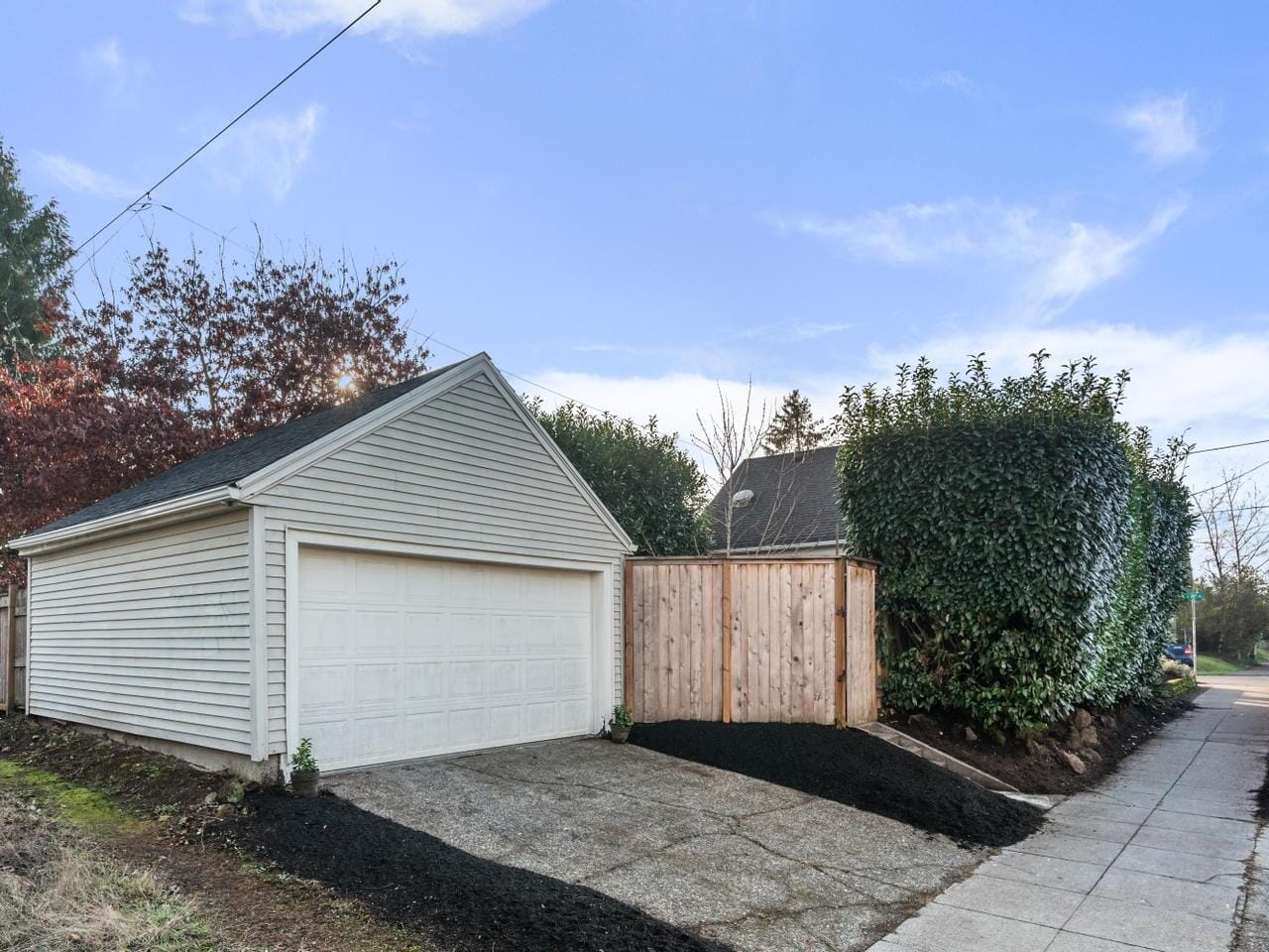 A small detached garage with a white door and beige siding is situated on a corner lot. It is surrounded by wooden fencing and greenery. The driveway leads to a sidewalk, and the sky is clear and blue.