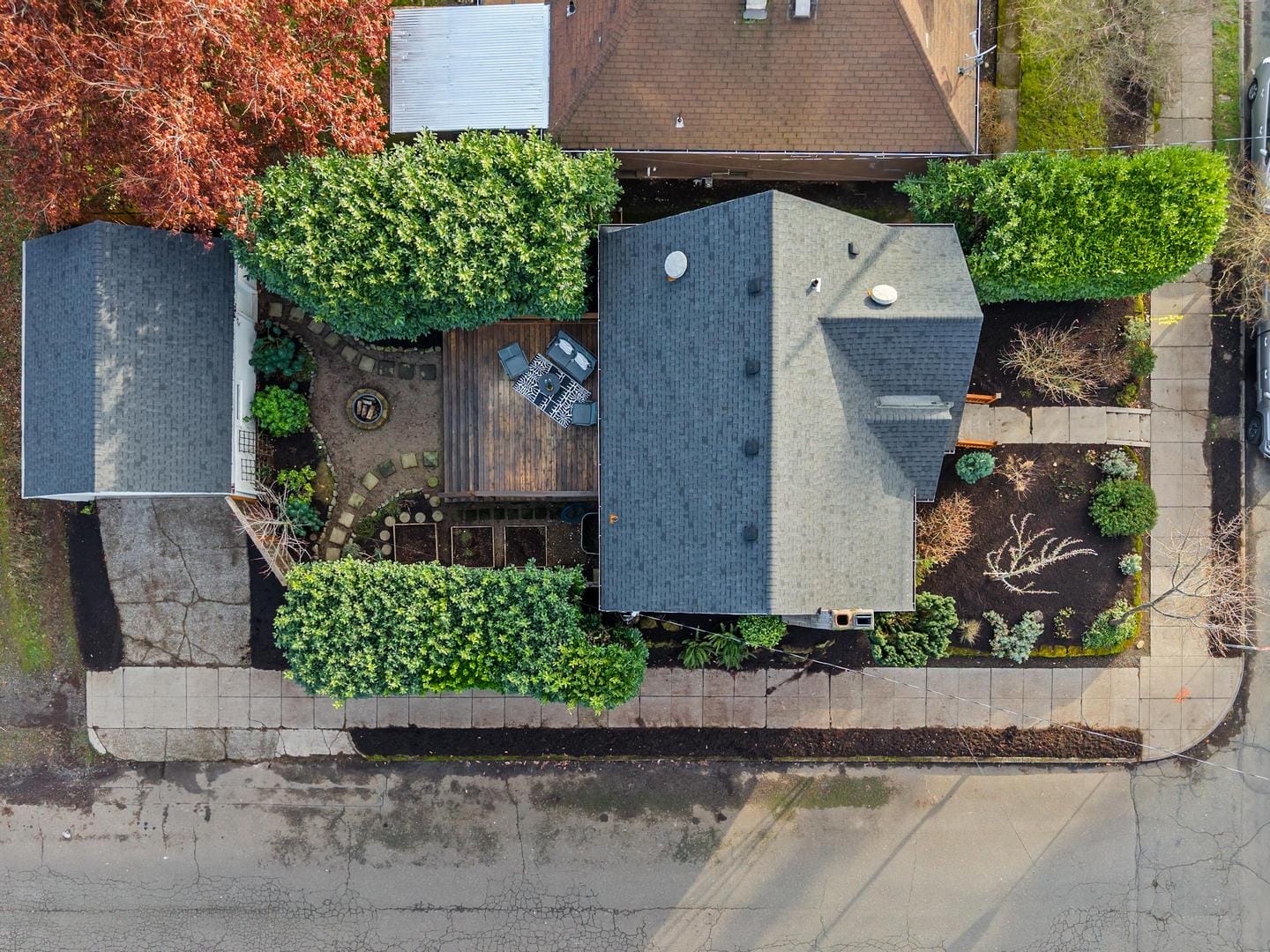 Aerial view of a suburban house with a gray roof, surrounded by green trees and shrubs. The backyard features a wooden deck with patio furniture and garden beds. The property is enclosed by sidewalks on three sides.