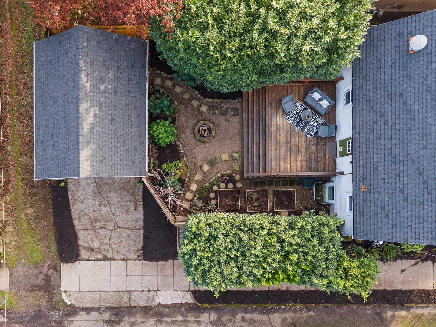 Aerial view of a cozy backyard with a wooden deck, outdoor furniture, and a small fire pit. The space is surrounded by lush greenery, a paved driveway, and a sidewalk. Raised garden beds are arranged near the deck.