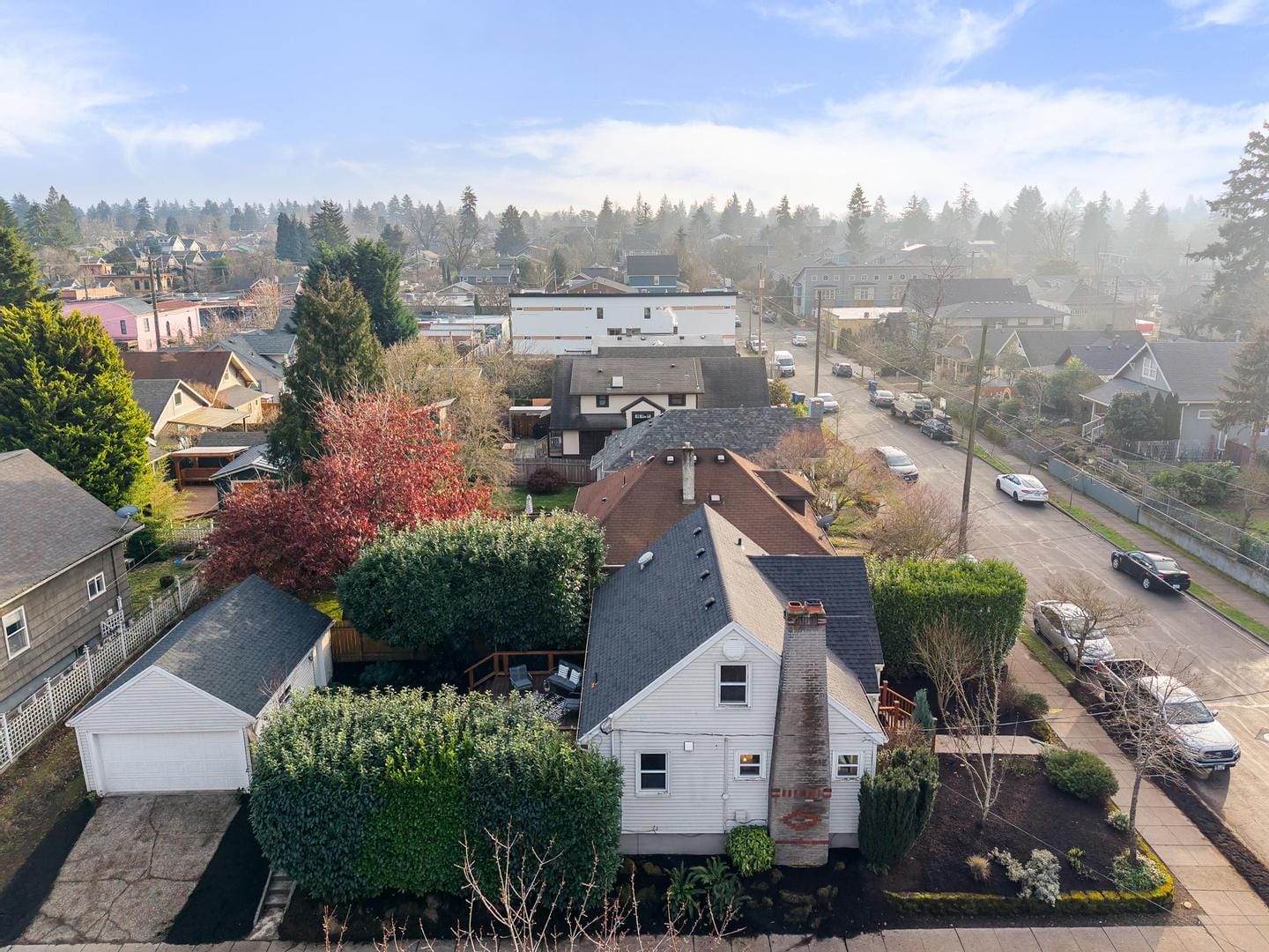 Aerial view of a suburban neighborhood with houses, trees, and a few cars parked on the street. The sky is clear with a few clouds, and the area is lush with greenery and landscaped yards.