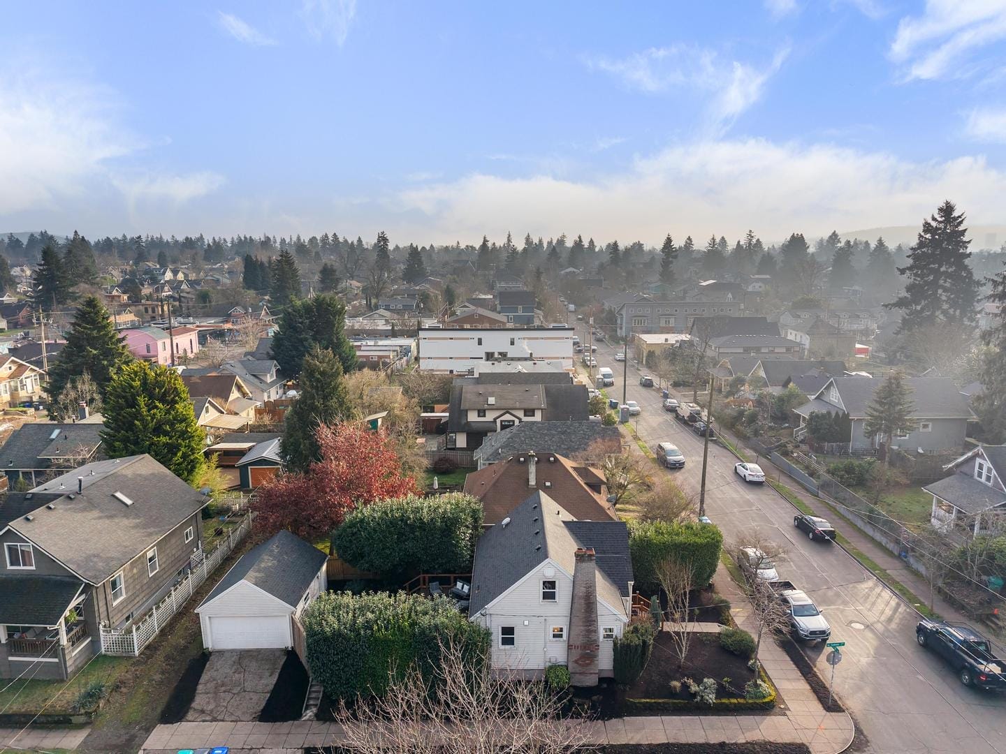 Aerial view of a residential neighborhood with houses surrounded by trees. A few streets intersect through the area, and a line of cars is parked along the road. The sky is clear with a few clouds.