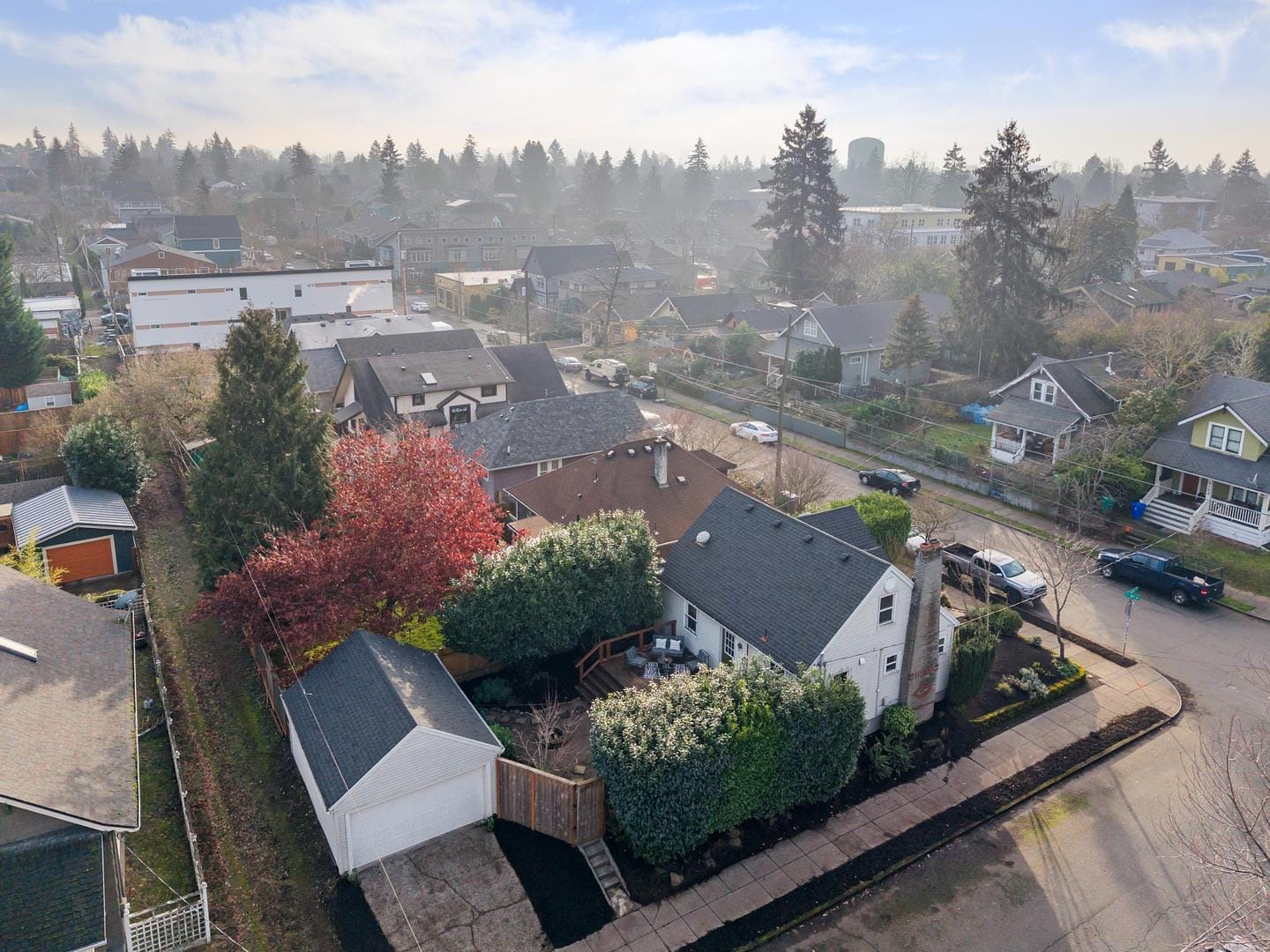 Aerial view of a suburban neighborhood with tree-lined streets and houses with varied roofs. A prominent tree with red leaves stands out in the center. The sky is clear, and the area is surrounded by distant trees and buildings.