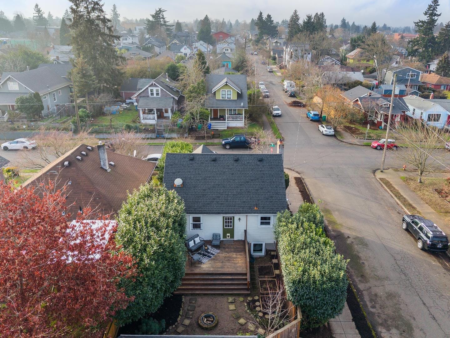 Aerial view of a residential neighborhood featuring a house with a dark roof and a wooden deck. Trees and a garden surround the home. The street has parked cars, and other houses with varied roof colors are visible in the background.