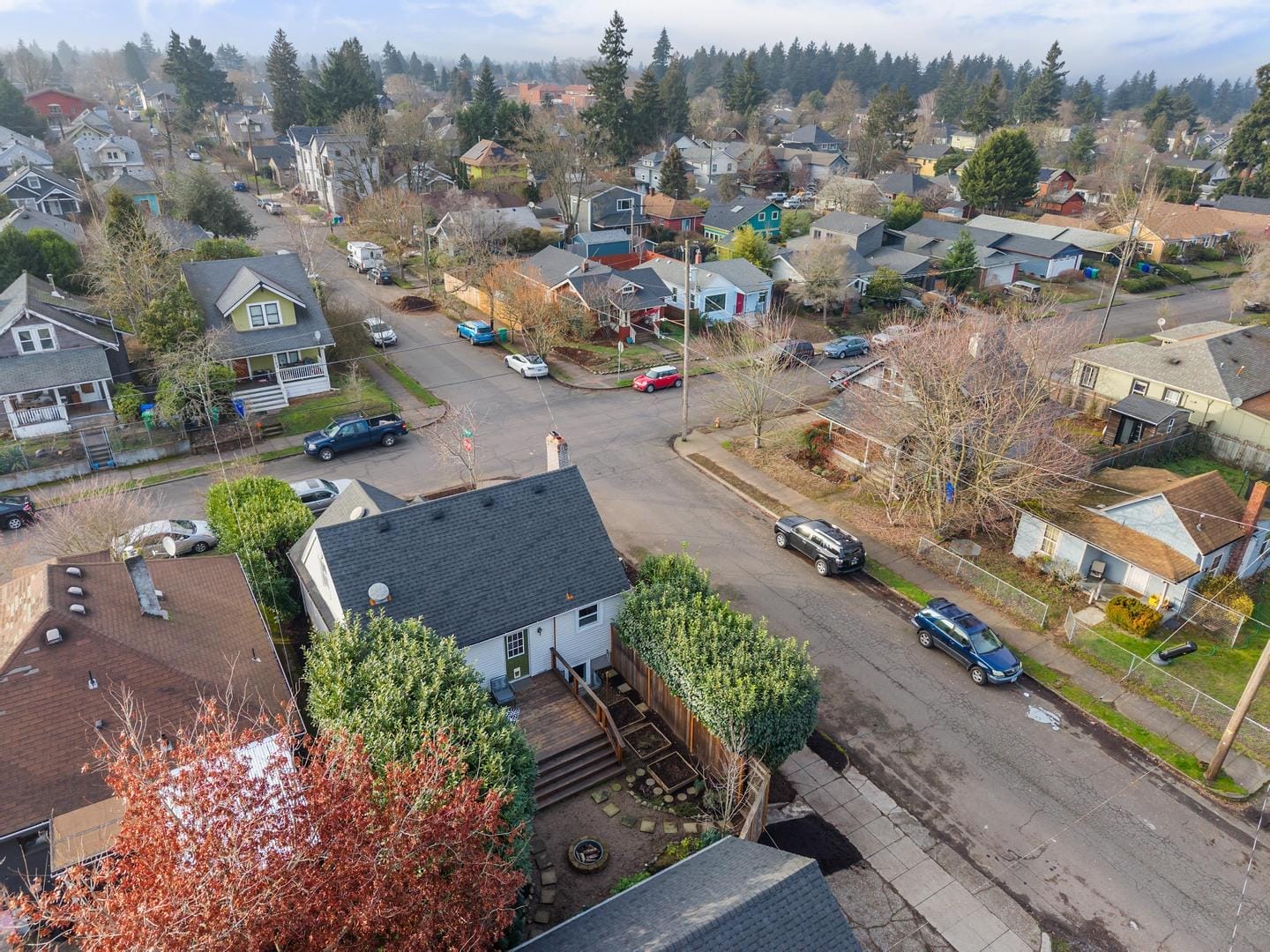 Aerial view of a suburban neighborhood with houses, trees, and parked cars. The streets form a grid with intersections, and the area is surrounded by greenery. The sky is partly cloudy.