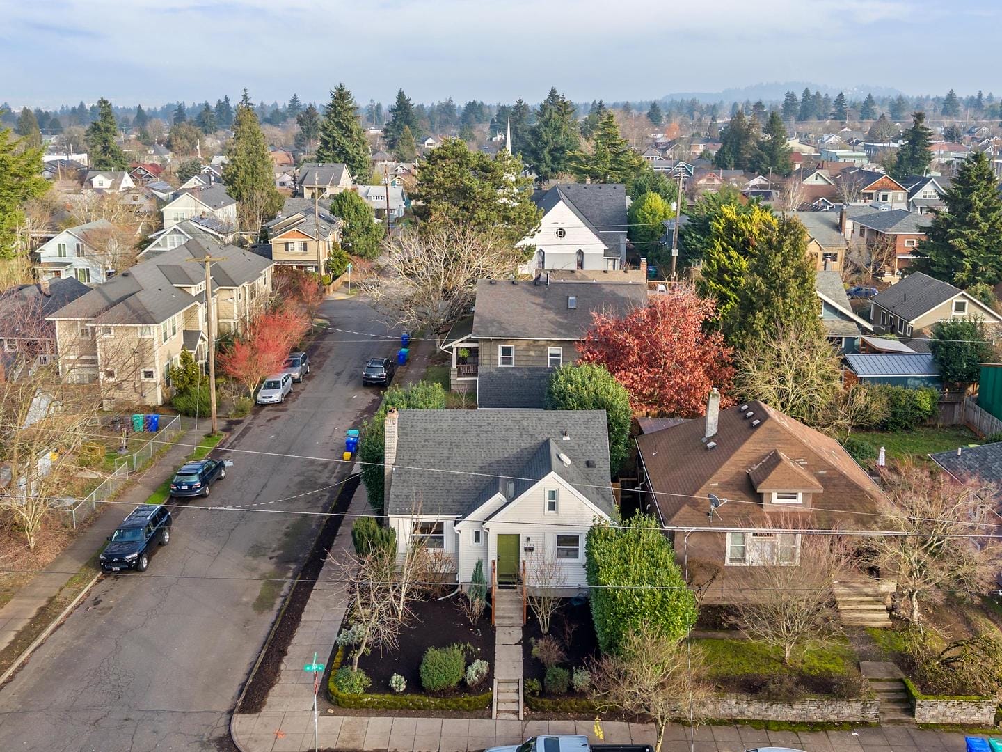 Aerial view of a suburban neighborhood featuring tree-lined streets and houses with various roof styles. The landscape includes small front yards, several cars parked along the street, and bare trees indicating a cooler season.