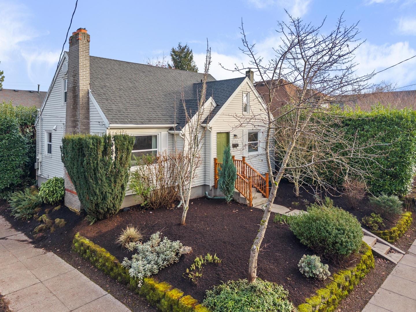 A small, well-maintained house with a gray roof and white siding. It has a green front door, surrounded by tidy landscaping, including shrubs and bare trees. The house is set against a clear blue sky, with a sidewalk and mossy curb lining the property.