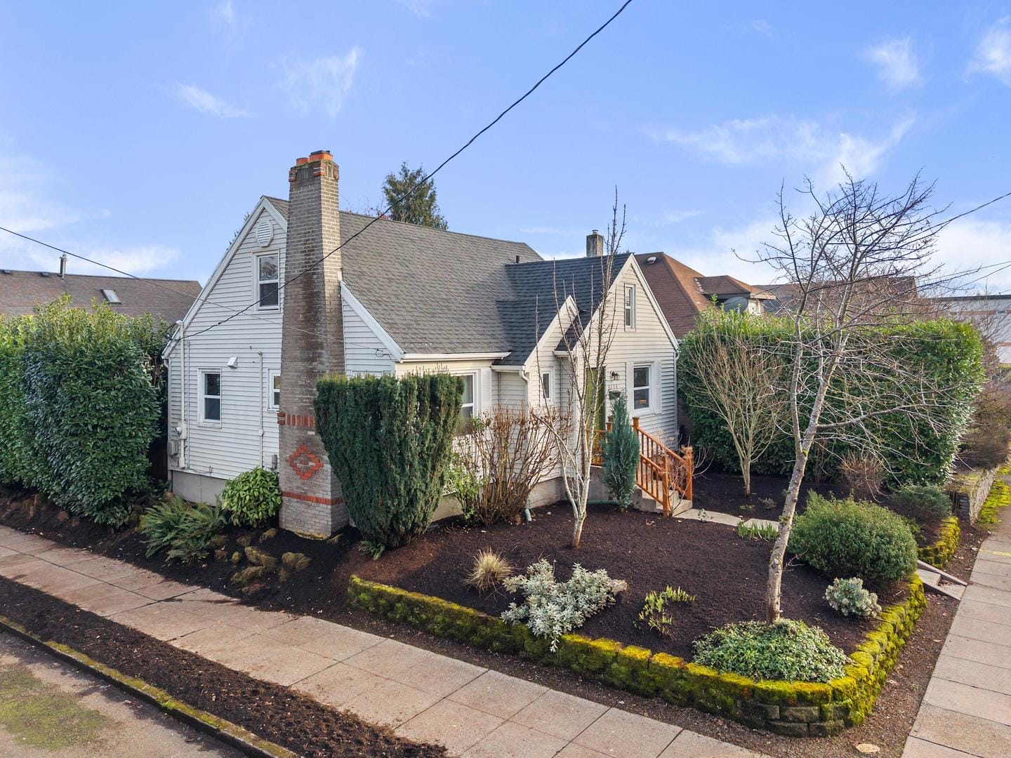 A small, single-story house with light siding and a gray roof features a brick chimney. It is surrounded by a neatly landscaped yard with shrubs and trees, bordered by a hedge. The sky is clear and blue.