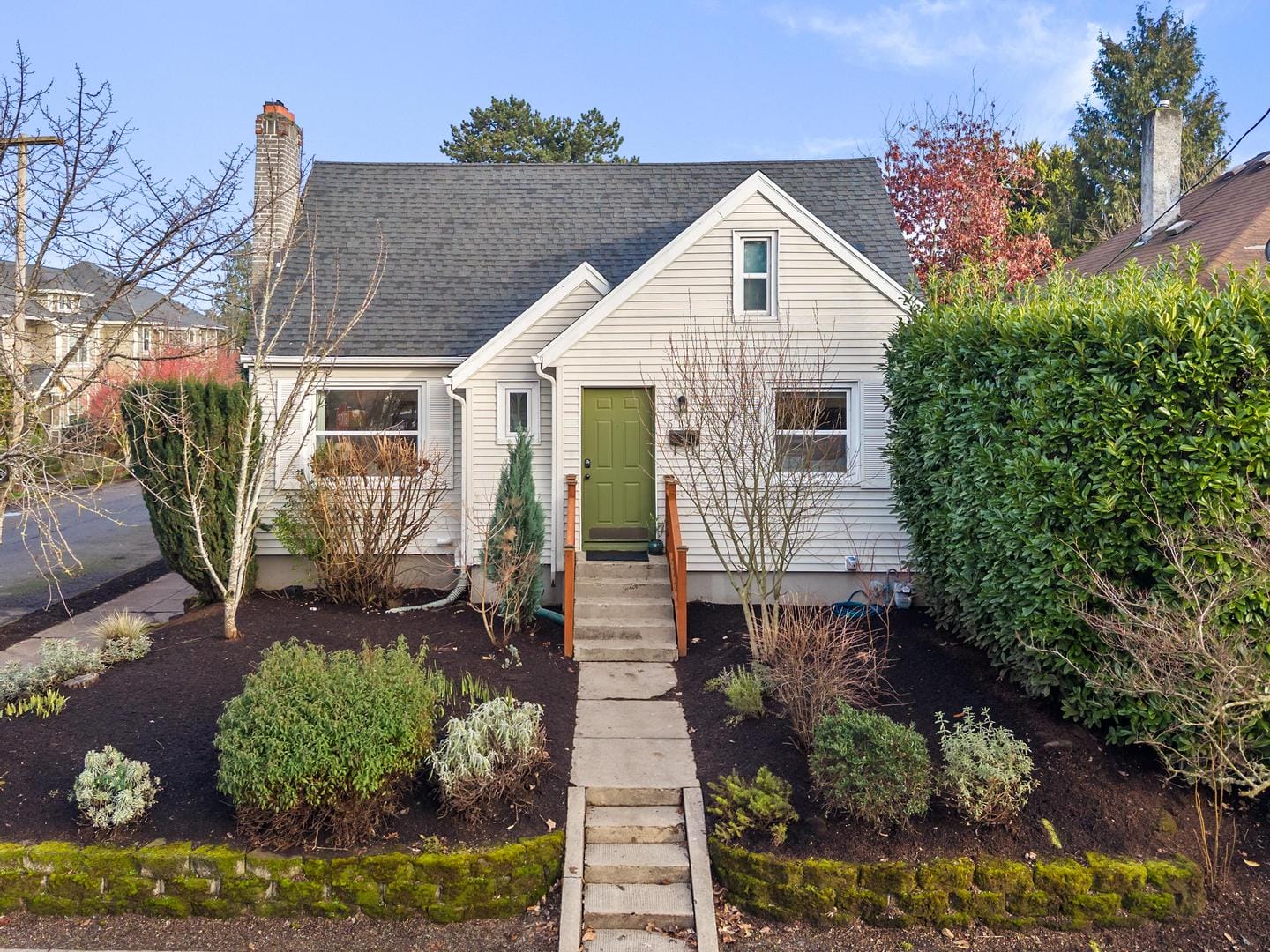 A small white house with a green front door, surrounded by neatly trimmed hedges and shrubs. Theres a stone pathway leading to the entrance, and trees frame the scene. The sky is clear and blue.