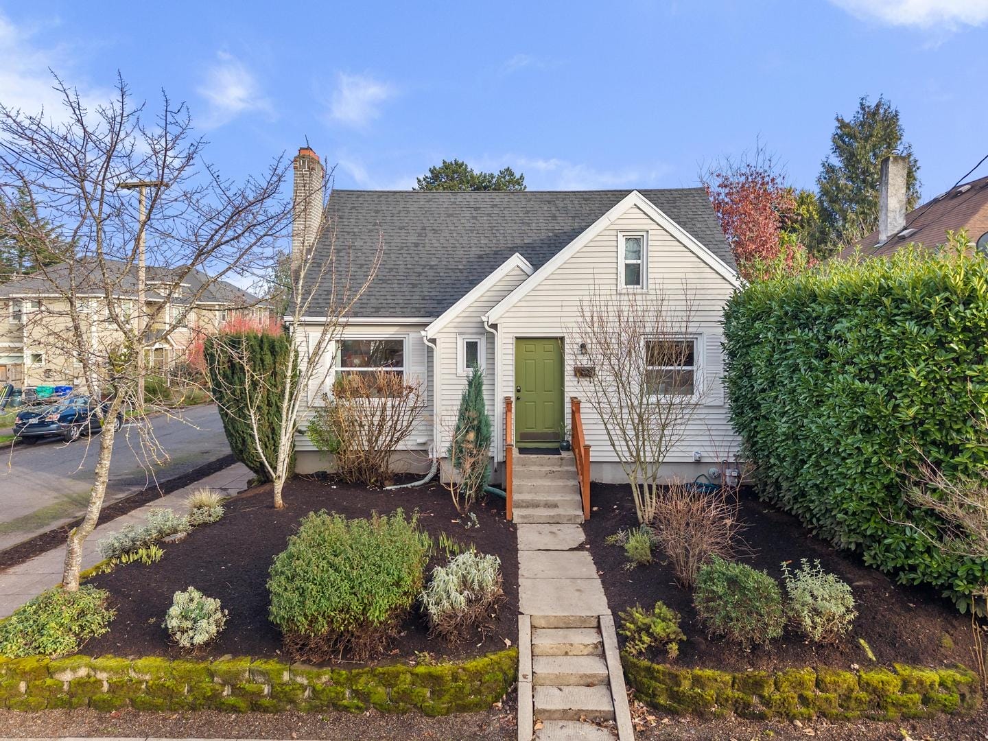 A charming, small white house with a green door and a neatly landscaped front yard. There are shrubs and small trees along a paved path leading to the entrance. The house is set against a blue sky.