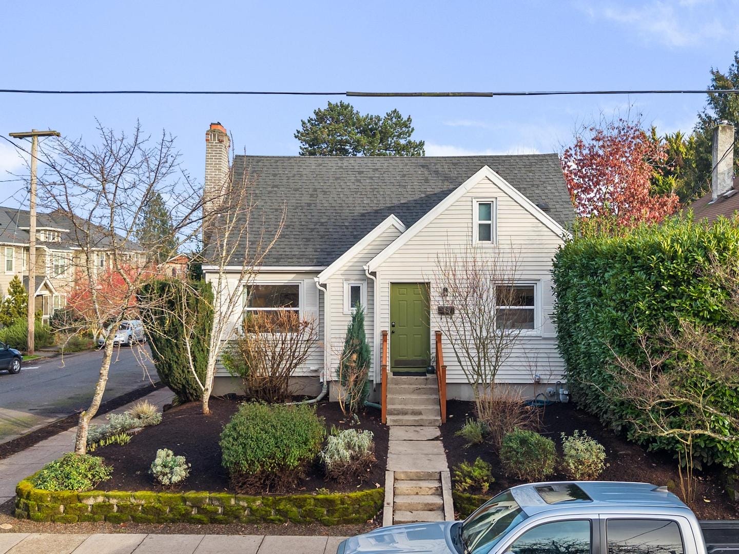 A small white house with a dark roof, green front door, and trimmed bushes in the front yard. A stone path leads to the porch. A car is parked on the street in front of the house, and bare trees frame the scene.