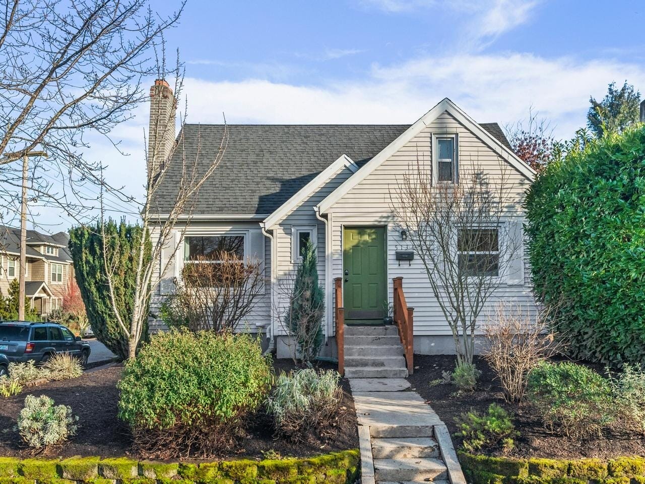 A charming, small white house with a dark roof and green front door. It has a stone pathway leading up to the entrance, surrounded by well-manicured shrubs and trees. The sky is clear and blue.