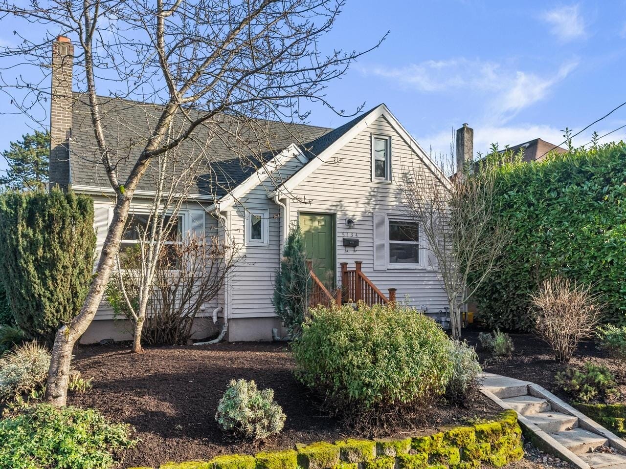 A quaint, single-story house with gray siding and a large dormer window. The entrance features a green door, and the yard is landscaped with shrubs and a leafless tree. A stone path leads to the door, bordered by a moss-covered edge.