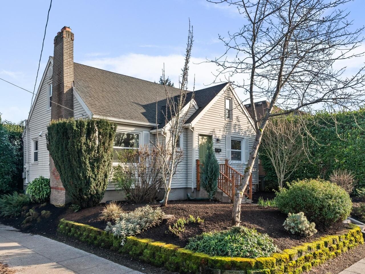 A charming, single-story white house with a steep gray roof and a brick chimney. Its surrounded by lush, well-maintained shrubs and trees, with a moss-covered stone wall bordering the front garden. A walkway leads to the front door.