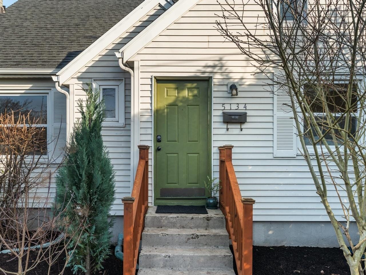 A white house with a green front door and house number 5134. The entrance has brown wooden railings and a few steps leading up to the door. Leafless trees and shrubs are visible in the small front yard.