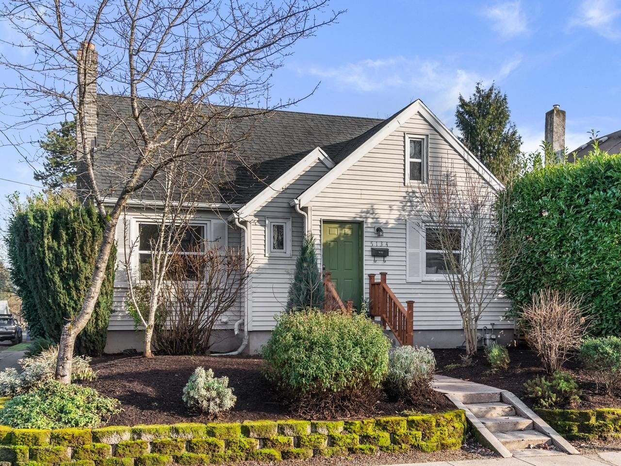 A charming small house with white wooden siding and a dark roof, surrounded by neatly trimmed bushes and trees. A short pathway leads to a green front door, and the garden is bordered by a low stone wall. Blue sky in the background.