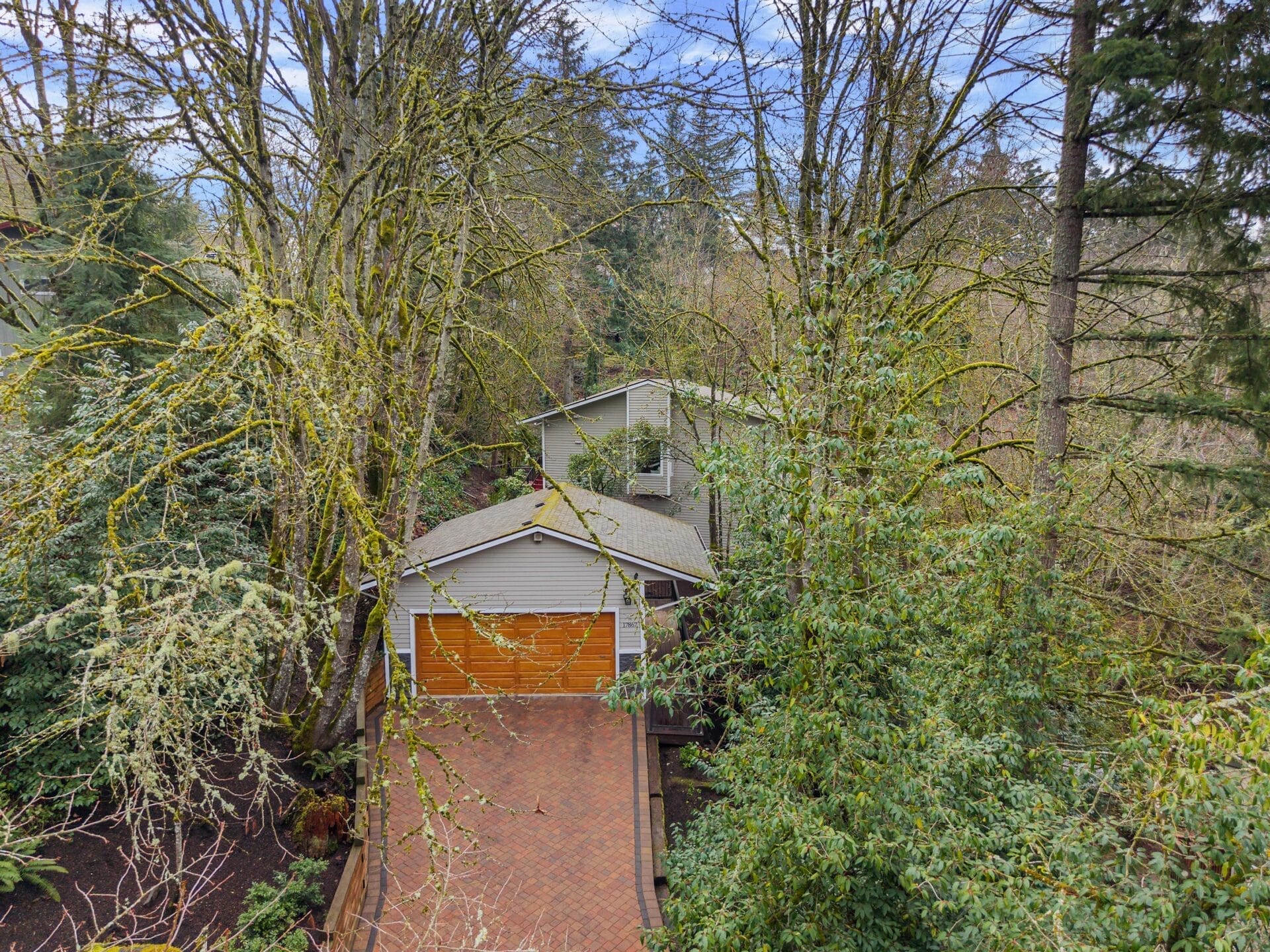 Aerial view of a two-story house surrounded by tall trees. The house has a gray exterior with a wooden garage door. A brick driveway leads to the entrance, and the area is lush with greenery, indicating a secluded, forested setting.