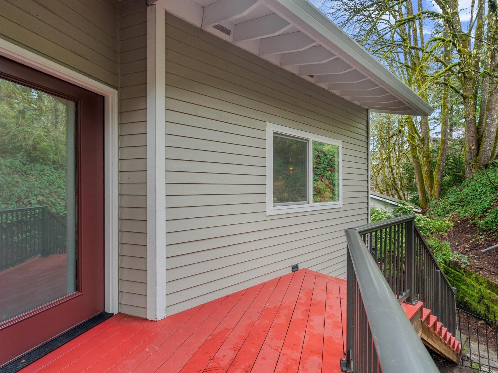 A small deck with red wooden flooring and gray siding on a house. It leads to a glass door on the left and a window in the center. The deck is bordered by a black railing and nearby trees provide a lush, green backdrop.