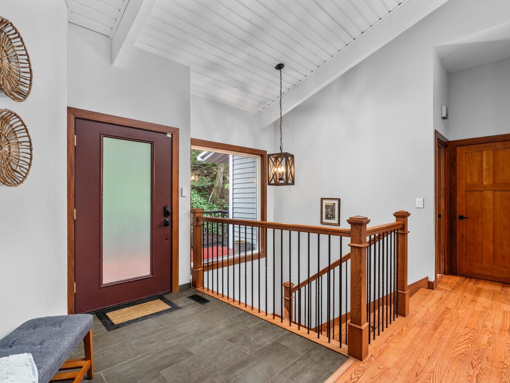 Spacious entryway with a modern design. Features a dark wooden front door with a frosted glass panel, a railing alongside stairs with wooden banisters, and a pendant light. A bench and woven wall decor are on one side, with hardwood floors throughout.