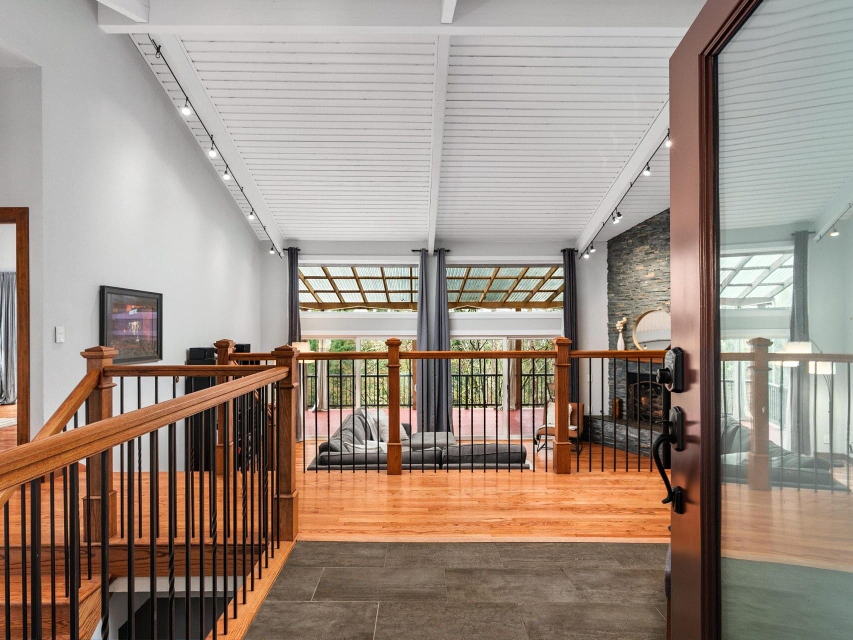 A modern indoor balcony with a slanted white ceiling and wooden floors. The space features dark iron railings, a seating area with a view of trees through a large window, and a stone wall with a fireplace. A glass door opens into the room.