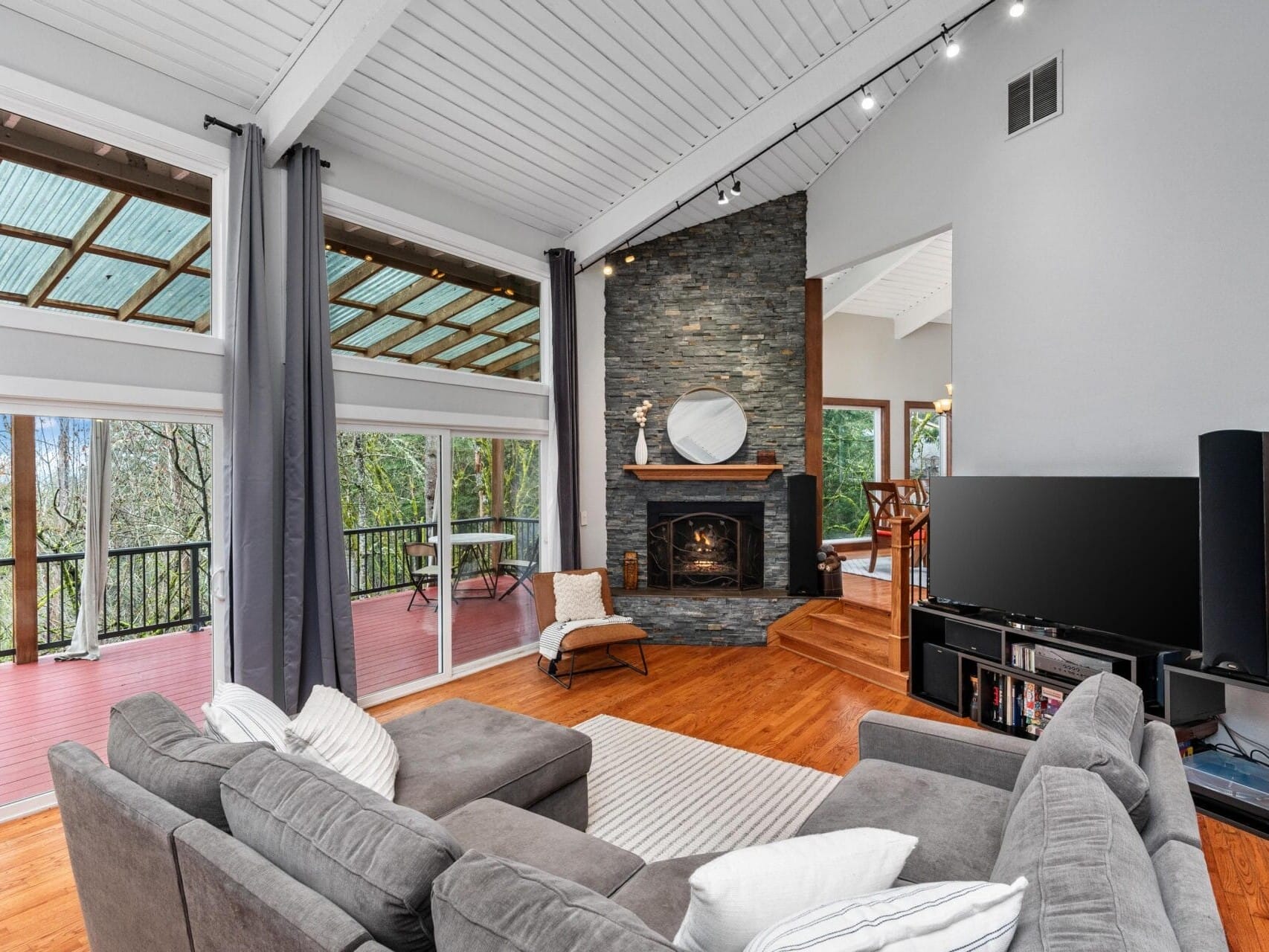 Spacious living room with gray sectional sofa, wooden floors, and a stone fireplace. Large windows and glass doors provide views of a red deck and trees. A flat-screen TV stands opposite the sofa, and a chair with a throw is near the fireplace.