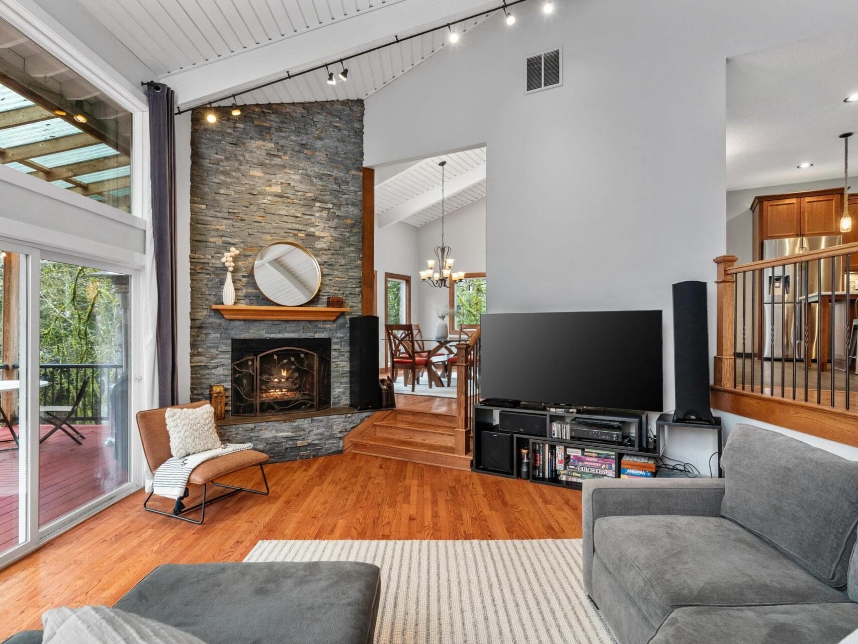 A modern living room with a stone fireplace, mirror above it, and a TV on a stand. Gray sofas are set on a wooden floor. A dining area with a chandelier is visible in the background. Large windows overlook a deck with outdoor seating.