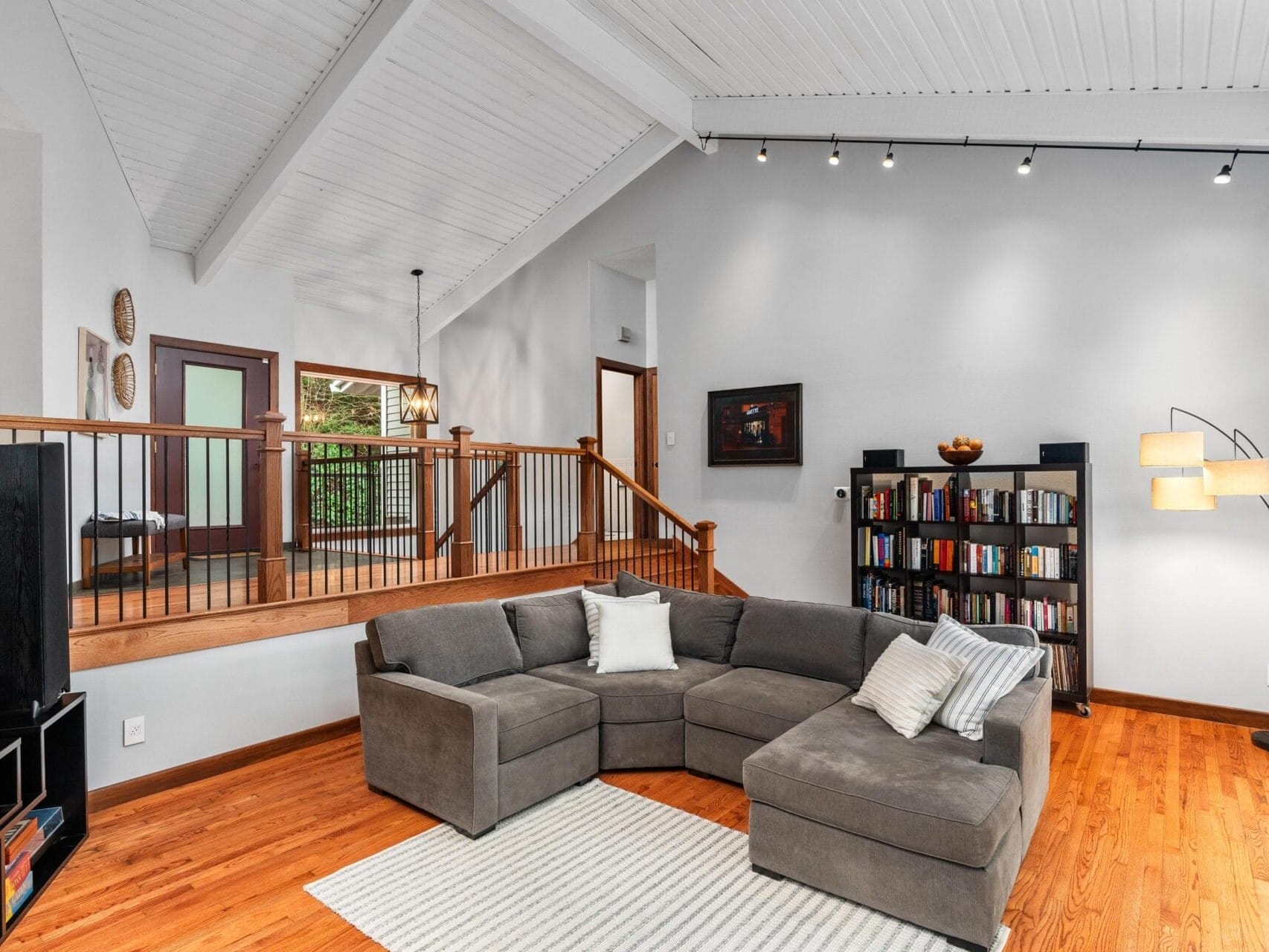 Spacious living room with a gray sectional sofa, white pillows, and a striped rug on wooden floors. Bookshelf against a pale wall with track lighting. Staircase with wooden railing leads to an upper level, and a floor lamp stands beside the sofa.