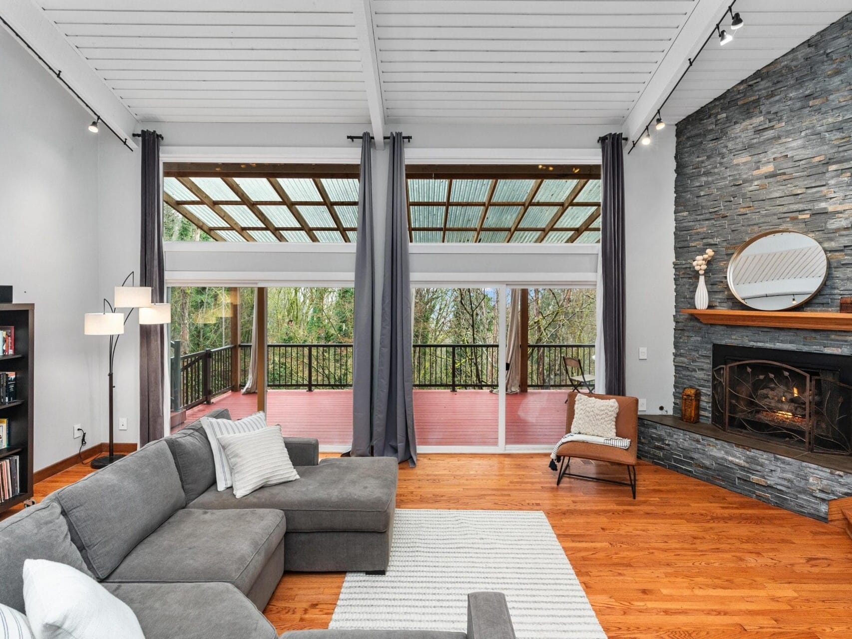 Spacious living room with a gray sectional sofa, stone fireplace, and large windows opening to a covered patio. The room features hardwood floors, a tall bookshelf, and decorative mirrors. White ceiling beams and hanging curtains add a modern touch.