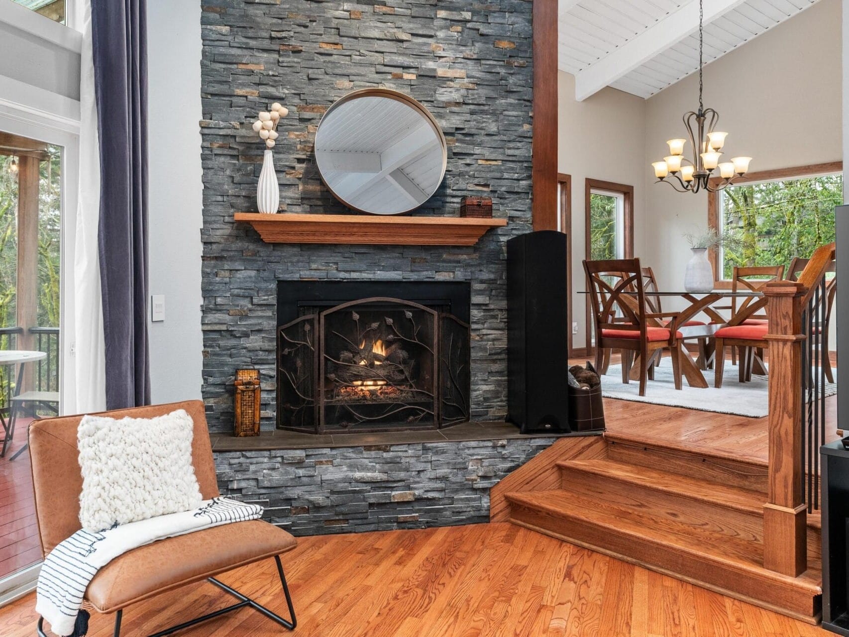 Cozy living room with a stone fireplace, wooden mantel, and round mirror above. A chair with a pillow sits nearby. Steps lead to a dining area with a chandelier and large windows showcasing a forest view. A deck is visible through the glass door.