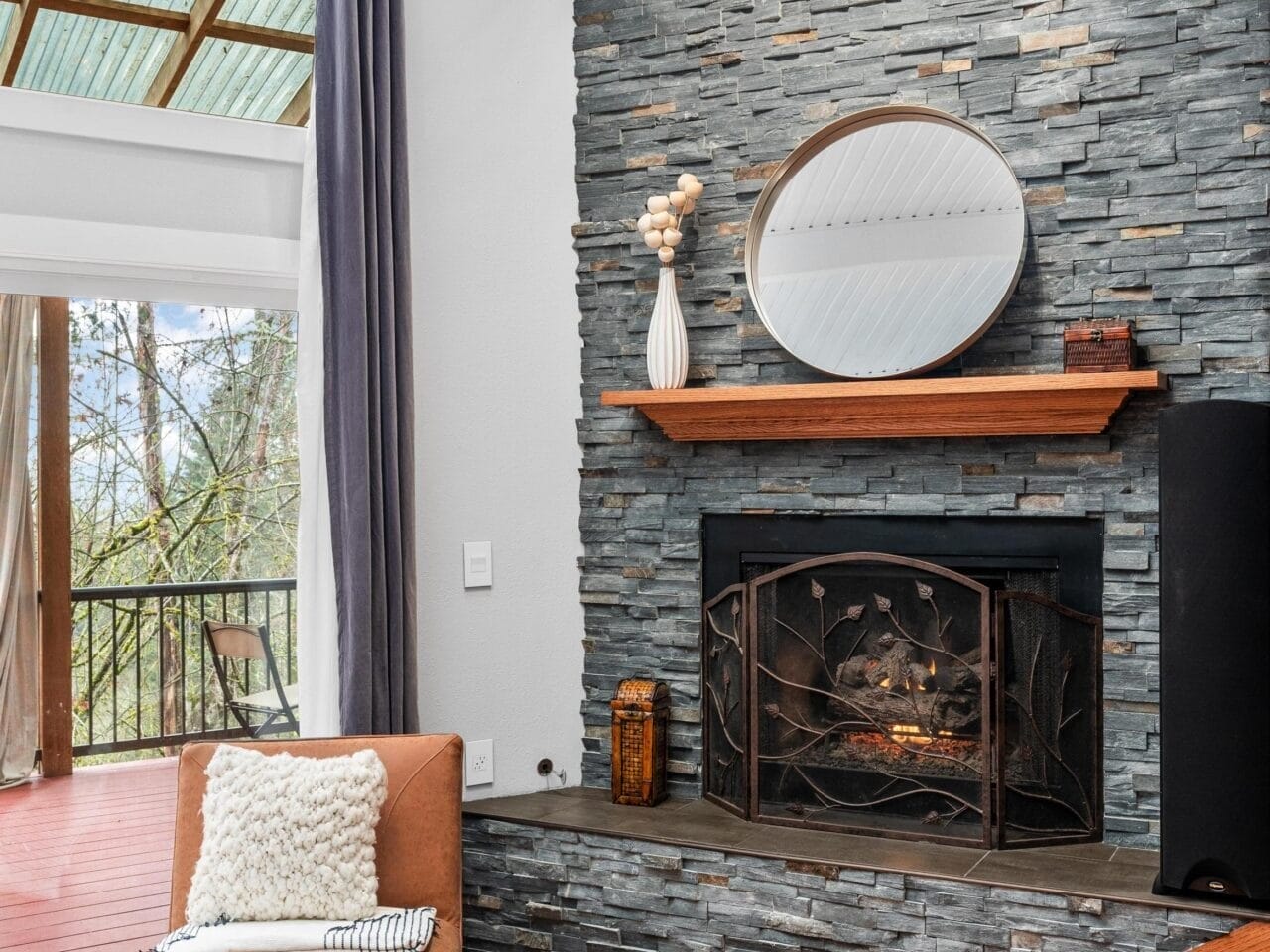 A cozy living room with a stone fireplace, round mirror, and decorative vase on the mantel. Theres a modern chair with a textured pillow nearby. The space has wooden flooring and large windows with a view of trees and a balcony outside.