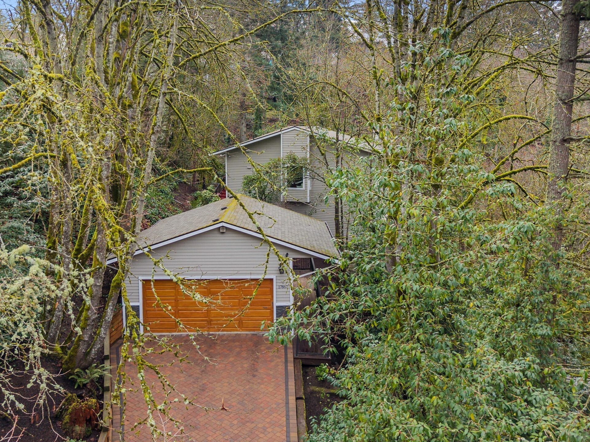 A house surrounded by dense trees with moss-covered branches. The two-story home has gray siding and a brown, double-door garage. The driveway is paved with red bricks, leading through leafy greenery in an overgrown, natural setting.