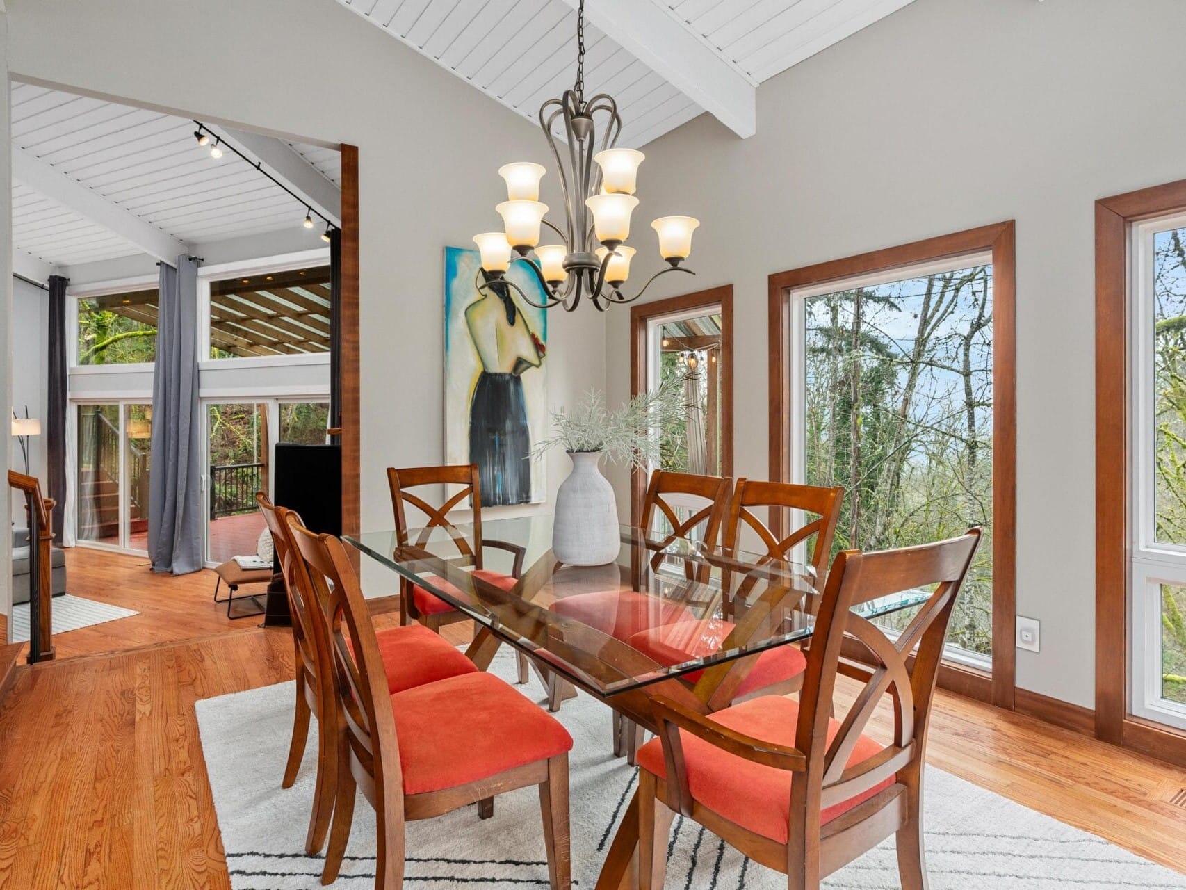 Dining room with a glass table and six chairs with red cushions. A modern chandelier hangs above. Large windows provide a view of trees outside. Light gray walls and wood floors create a warm atmosphere. An abstract painting and vase adorn the space.