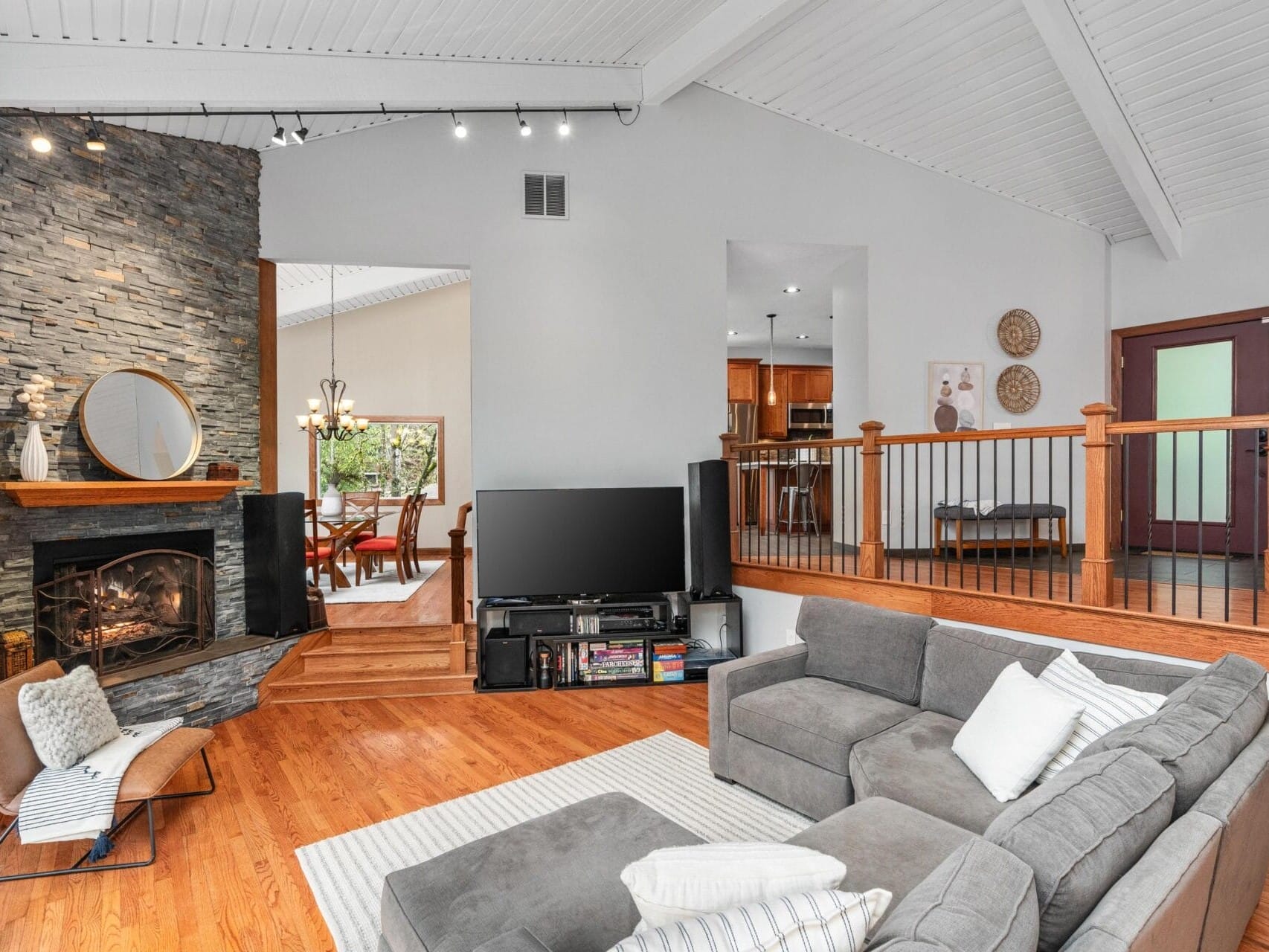 A modern living room with a gray sectional sofa, stone fireplace, and flat-screen TV. The room is bright with a high, white vaulted ceiling. A dining area is visible through a doorway, featuring a wooden table and a large window.