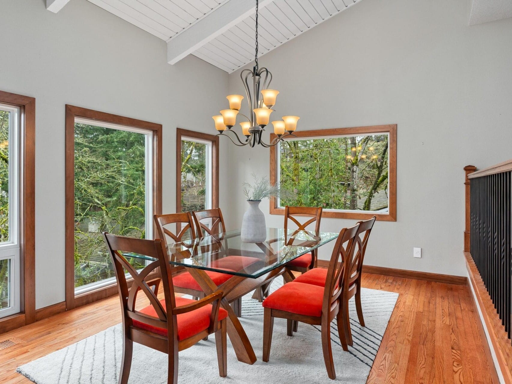 Dining room with a glass table and six wooden chairs with red cushions. A modern chandelier hangs above the table. Large windows offer a view of trees outside, and the room has wooden floors and neutral walls. A white vase sits on the table.