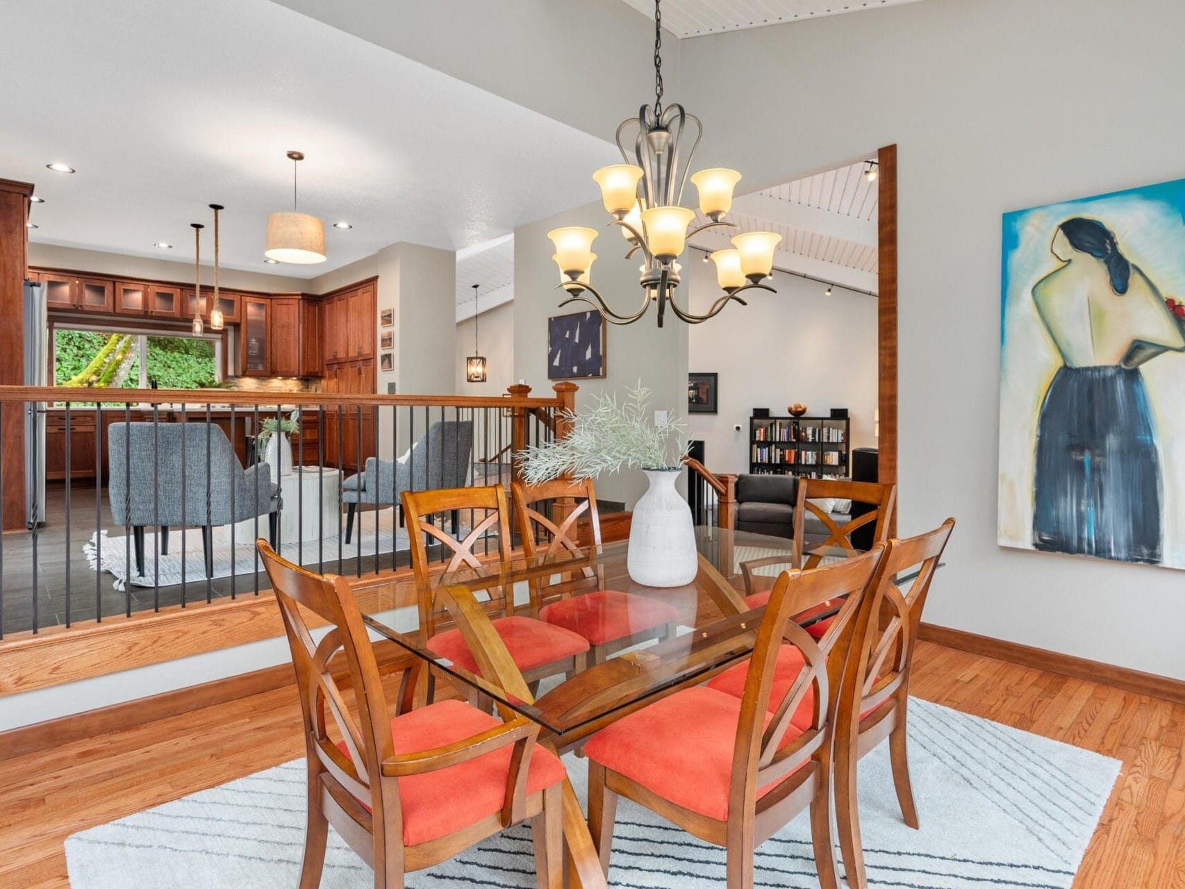 A dining area with a glass table and six wooden chairs with orange cushions. A modern chandelier hangs overhead. An abstract painting is on the wall. In the background, a kitchen and living area with gray chairs are visible. Wood flooring throughout.