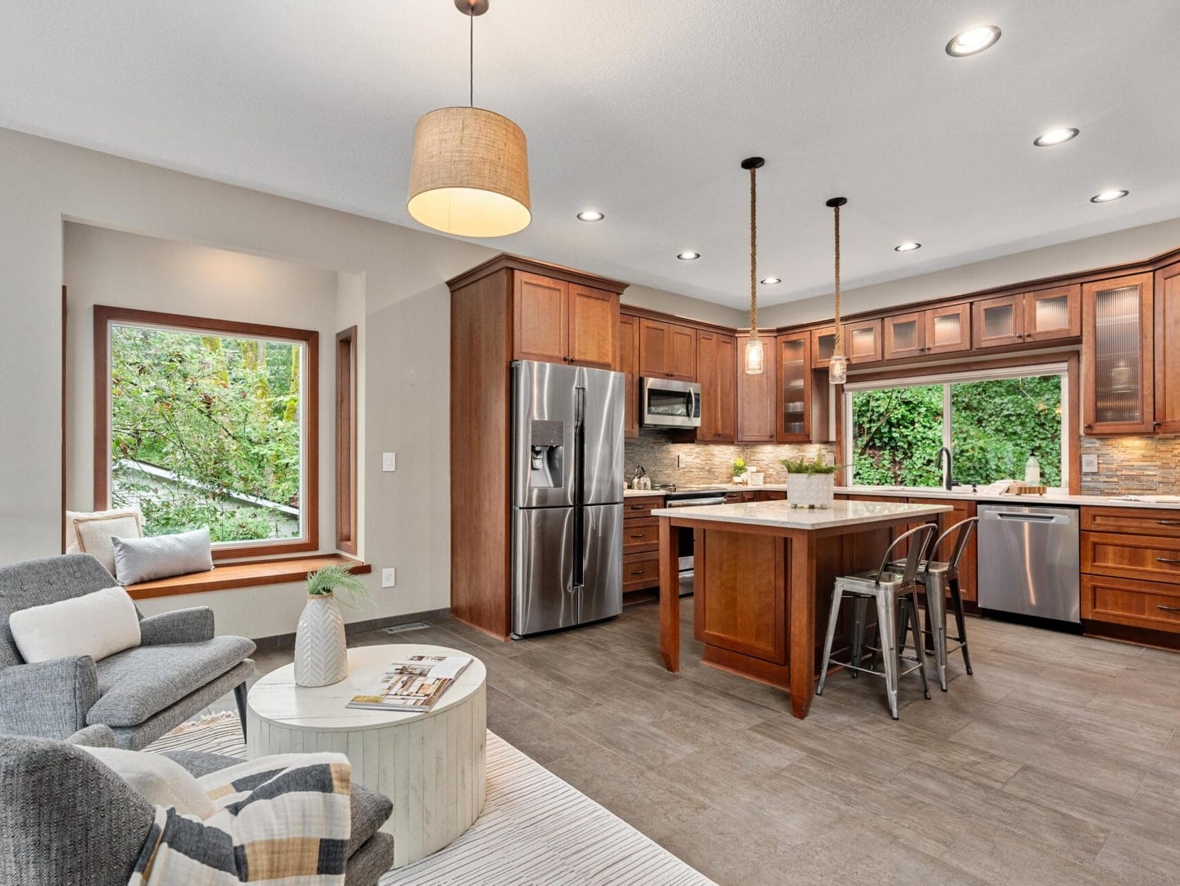 Modern kitchen with wooden cabinets, a central island, and stainless steel appliances, including a fridge and dishwasher. Two bar stools are at the island. To the left, theres a cozy seating area with gray chairs and a large window with a view of trees.