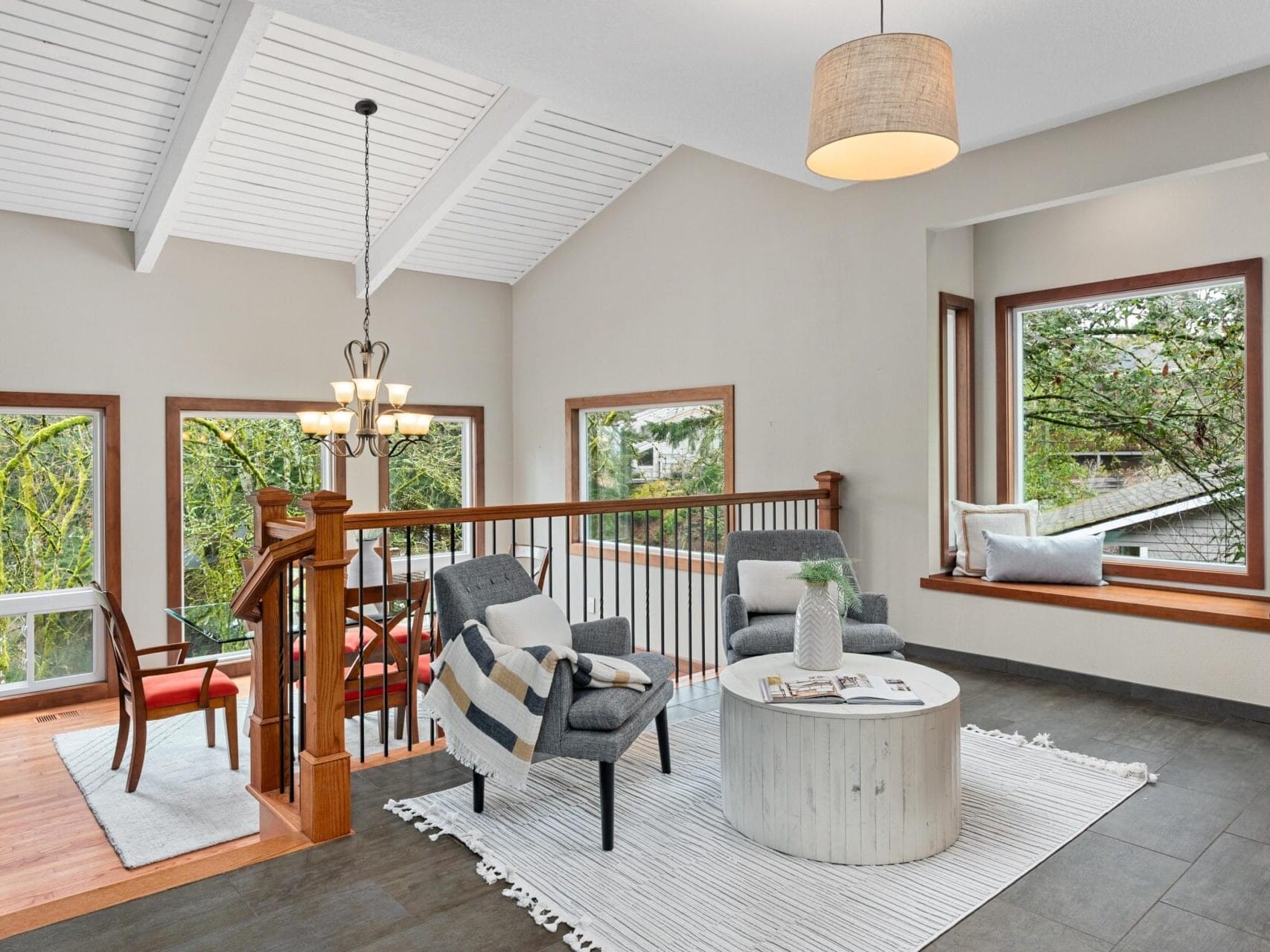 Modern living room with high ceiling and large windows displaying a forest view. Two armchairs and a round coffee table on a light rug face a staircase with wooden railing. A hanging lamp and dining area with red chairs are visible in the background.