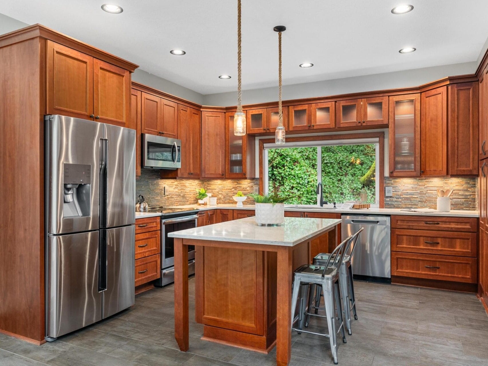 Modern kitchen with wooden cabinets, stainless steel appliances, and a central island with a white countertop. Hanging pendant lights illuminate the space. Large window with a view of greenery. Gray floor tiles and an overall warm ambiance.