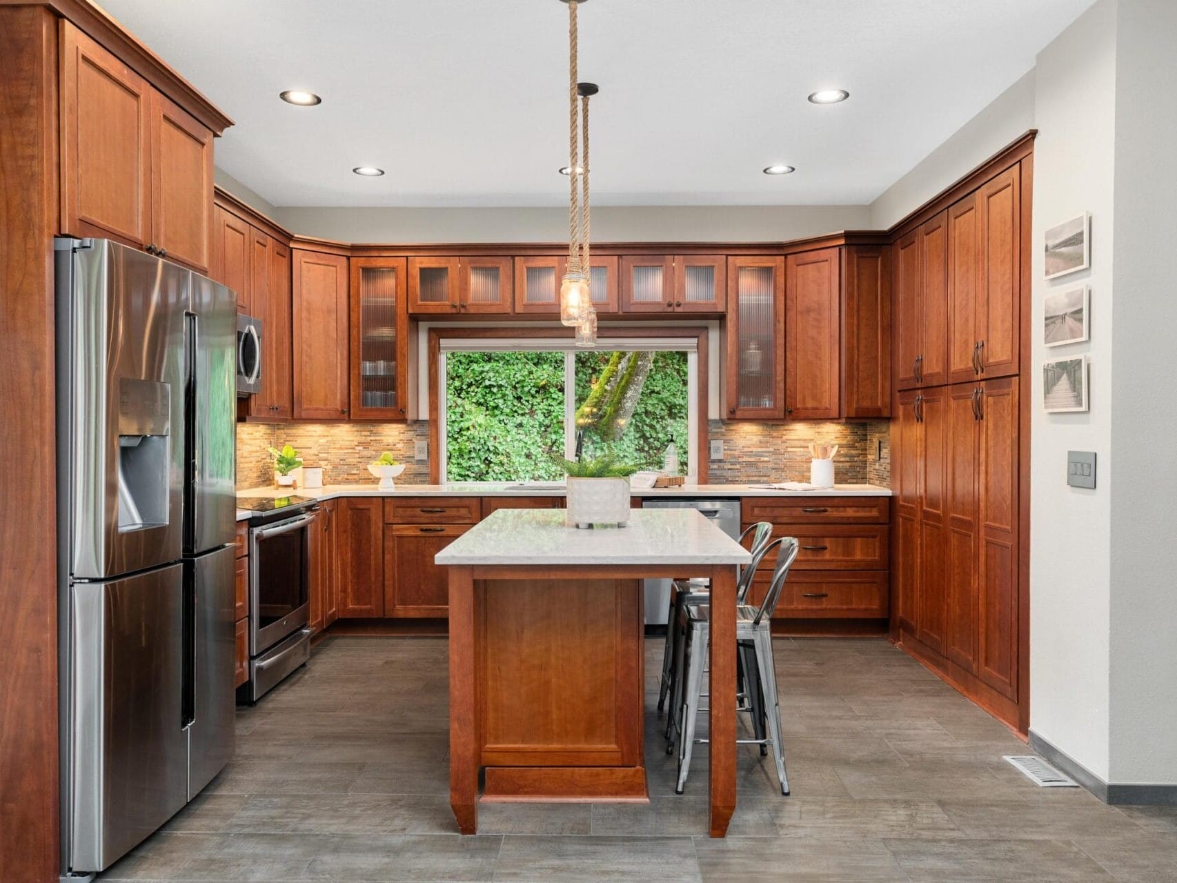 A modern kitchen with wooden cabinets and a central island featuring a white countertop. Stainless steel appliances, including a fridge and oven, are present. Pendant lighting hangs above the island, and a large window provides a view of greenery outside.