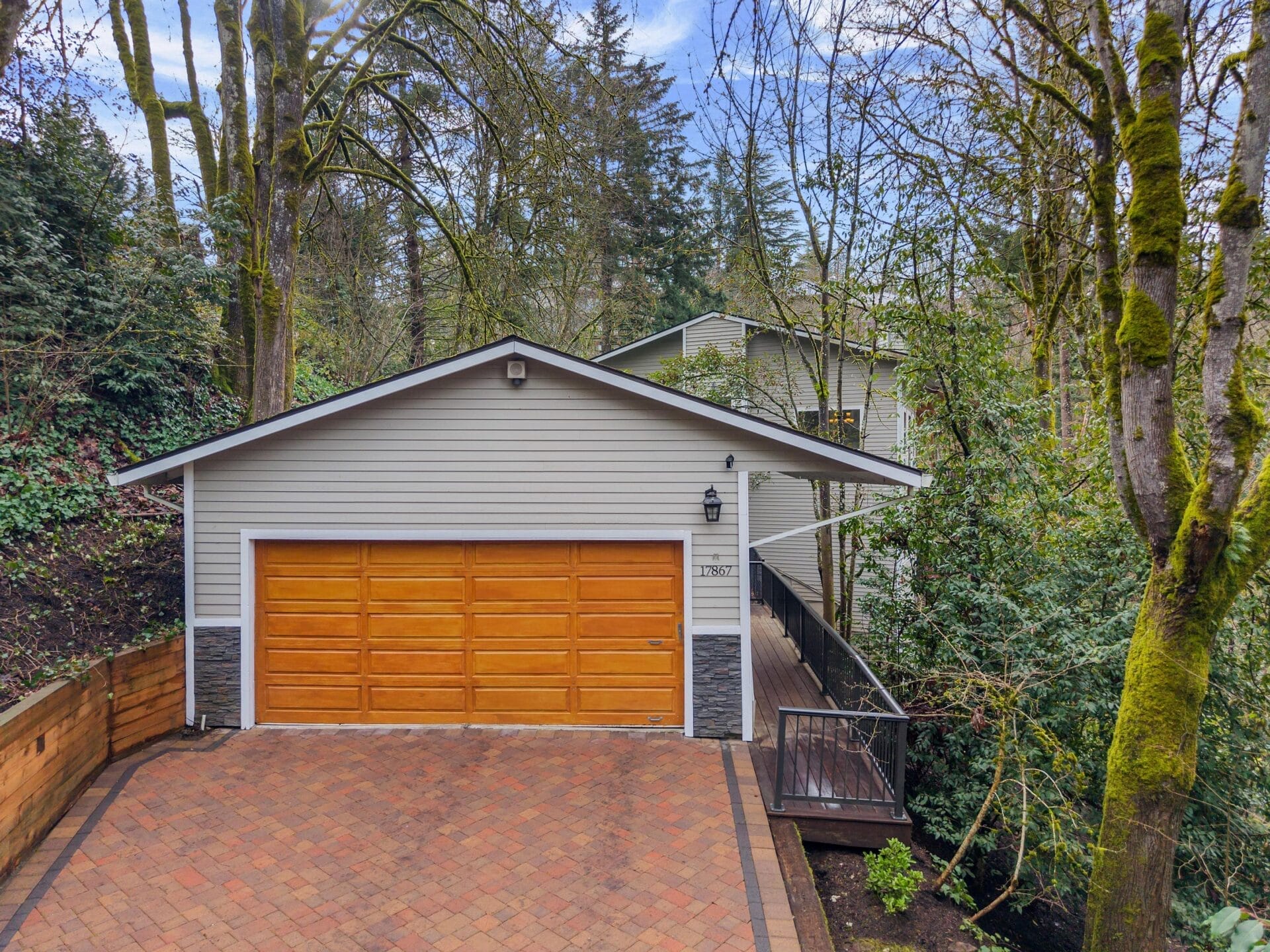 A modern two-story house surrounded by trees, featuring a light gray exterior with a wooden double garage door. A pathway leads to the entrance on the right, and the driveway is paved with red-brown bricks.