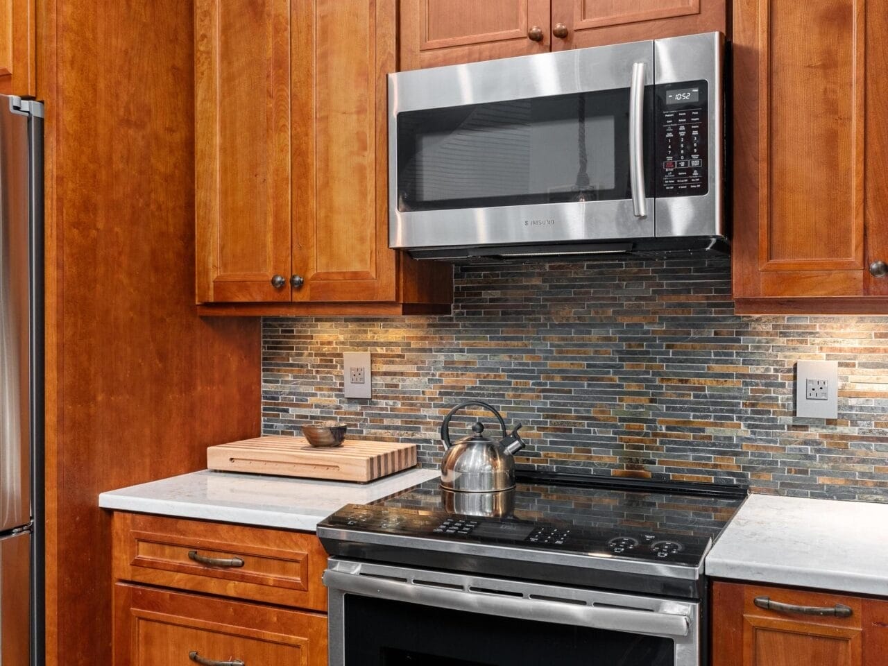 A kitchen with wooden cabinets, a stainless steel oven, microwave, and refrigerator. The backsplash features a mosaic tile design. A silver kettle sits on the stove, and a wooden cutting board is on the counter.